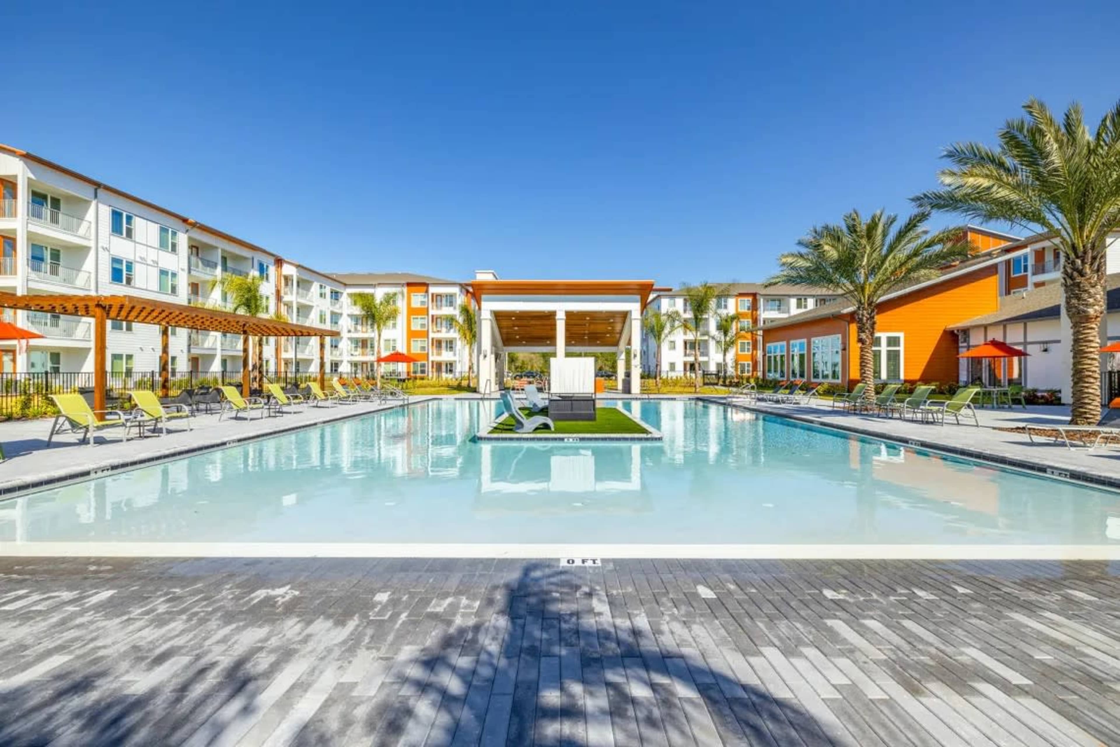 A large swimming pool is surrounded by lounge chairs and palm trees, with a modern clubhouse in the background.