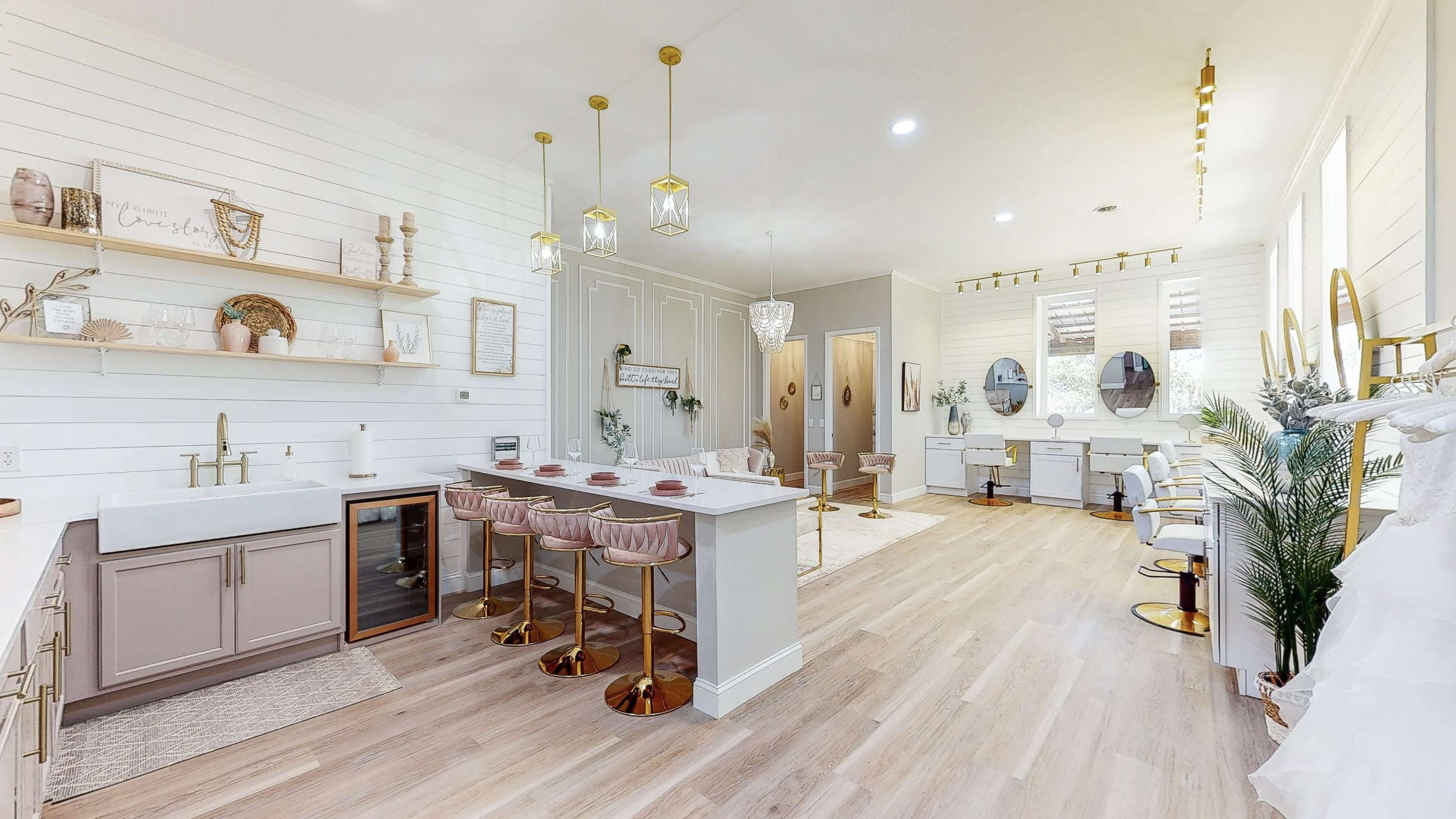A spacious, modern salon with light wood flooring, a kitchen-like area with bar seating, and multiple styling stations featuring mirrors and chairs.