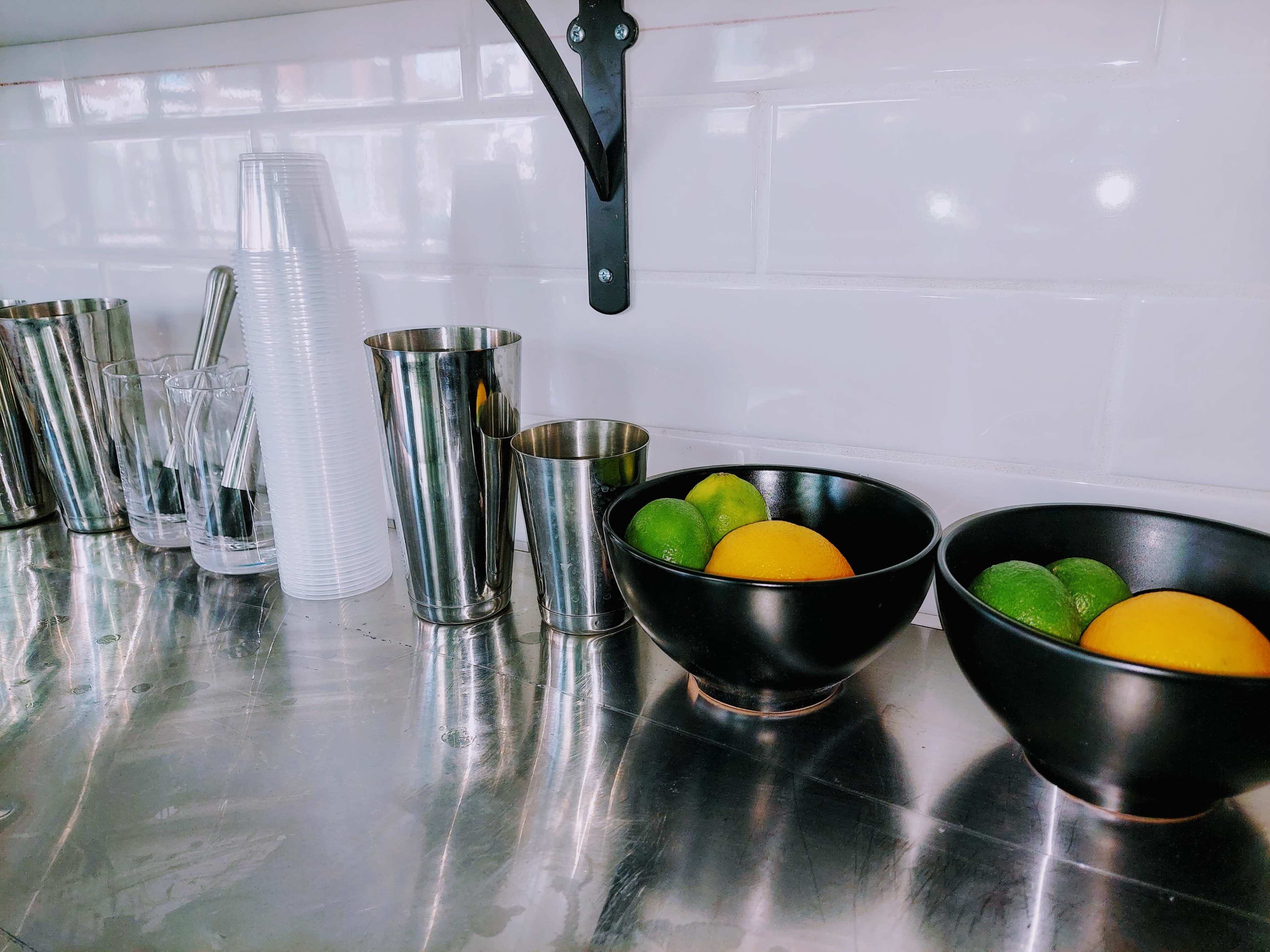 A countertop with various metallic cups and glasses alongside two bowls containing oranges and limes.