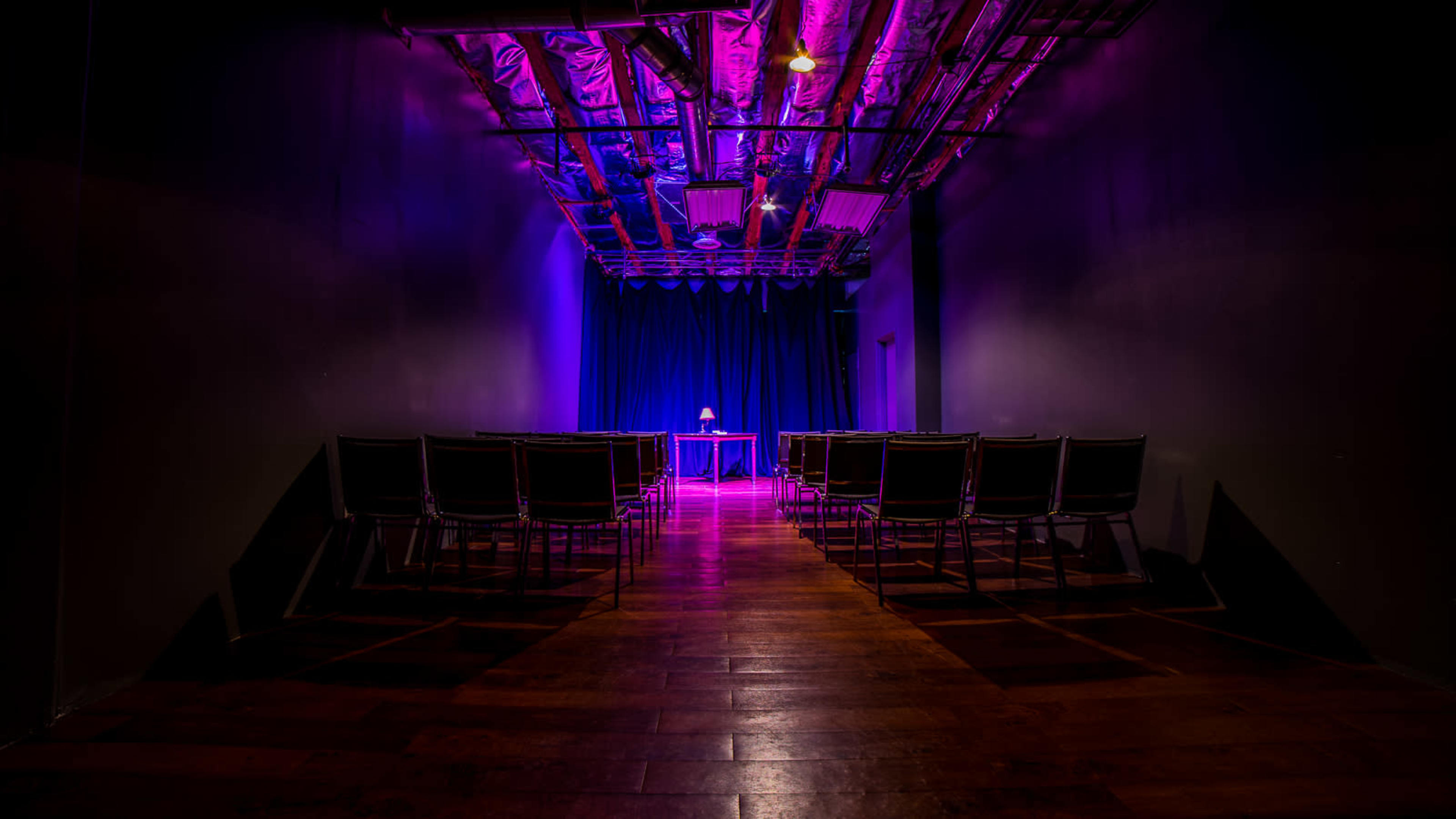 A dimly lit performance space features rows of chairs facing a small table illuminated by a purple light.