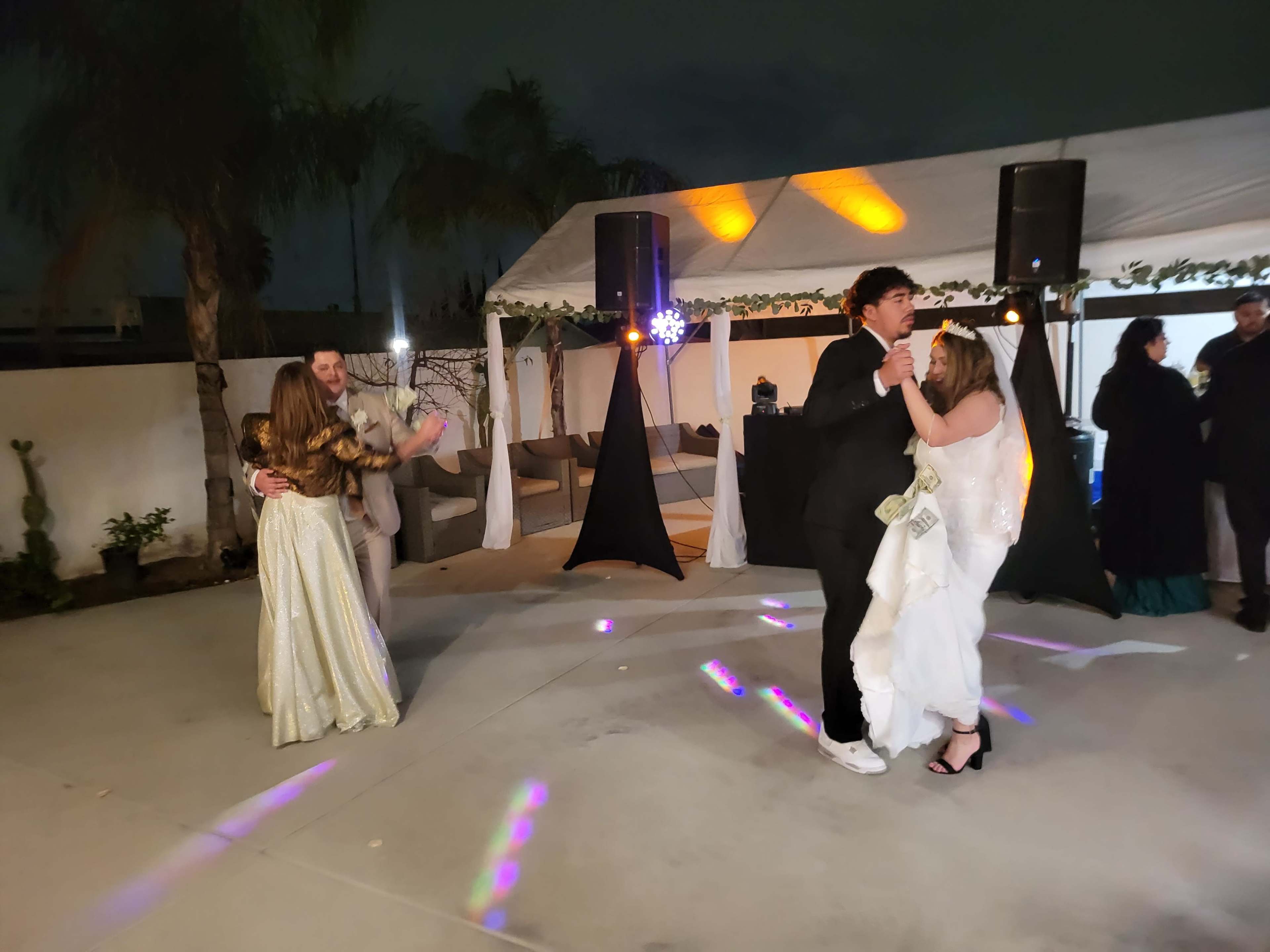 A group of four people dances under festive lighting at an outdoor wedding reception.