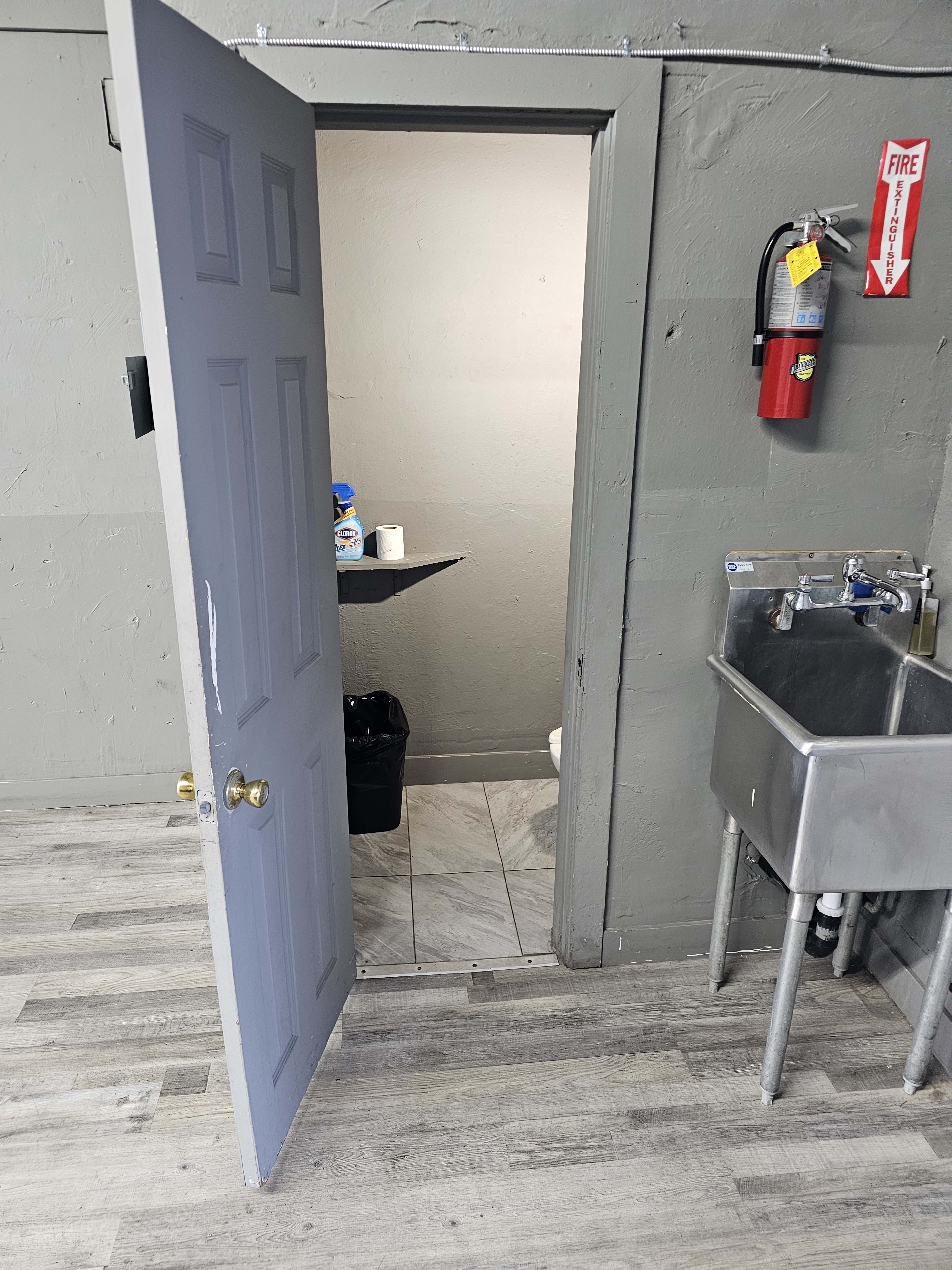 A gray door stands open, revealing a small restroom with a toilet and a sink adjacent to a utility sink in a room with light-colored flooring.