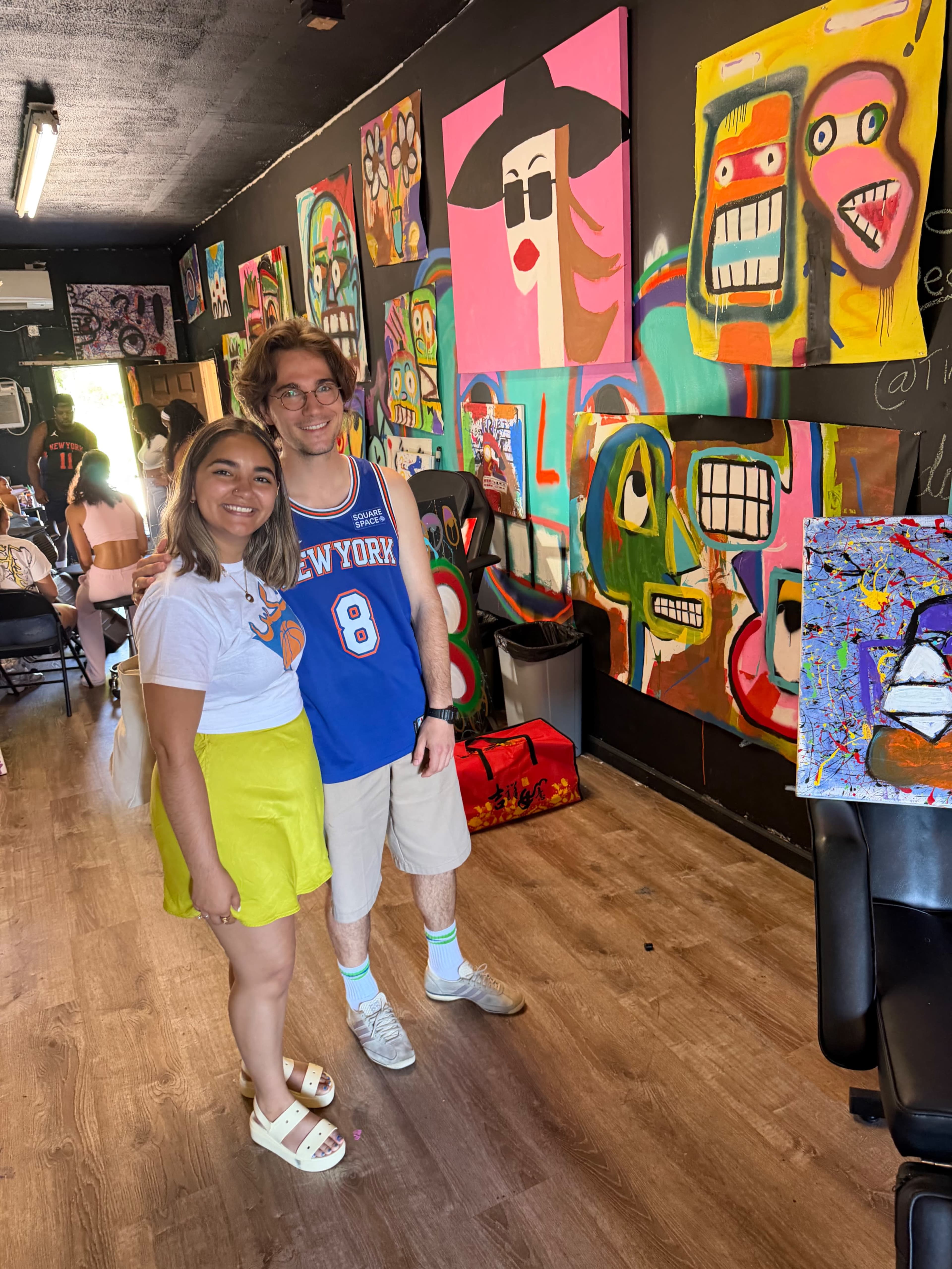 Two people are posing together in an art studio filled with colorful paintings.