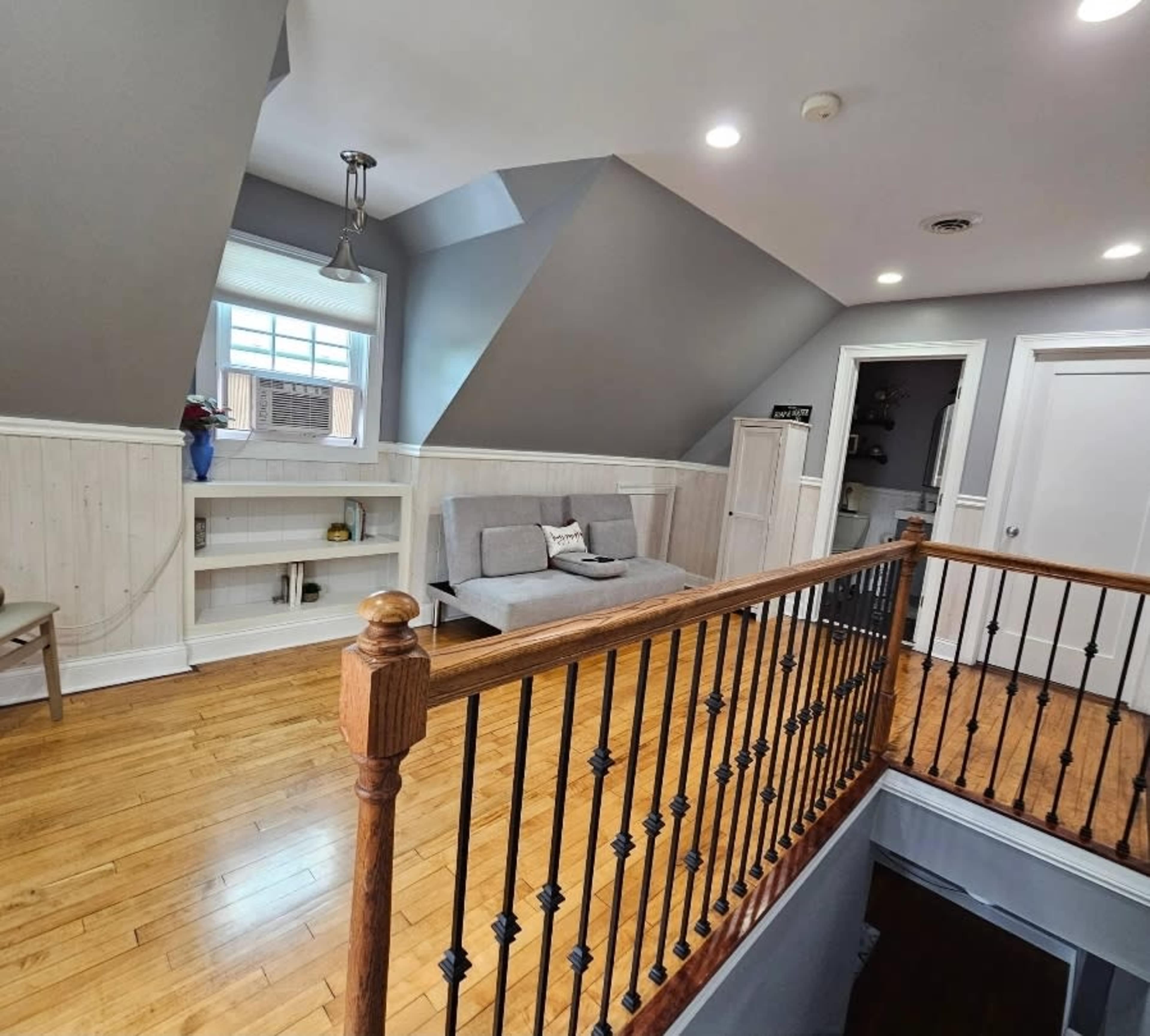 The image shows a loft space with sloped ceilings, hardwood floors, a gray couch, and a railing leading to an open staircase.