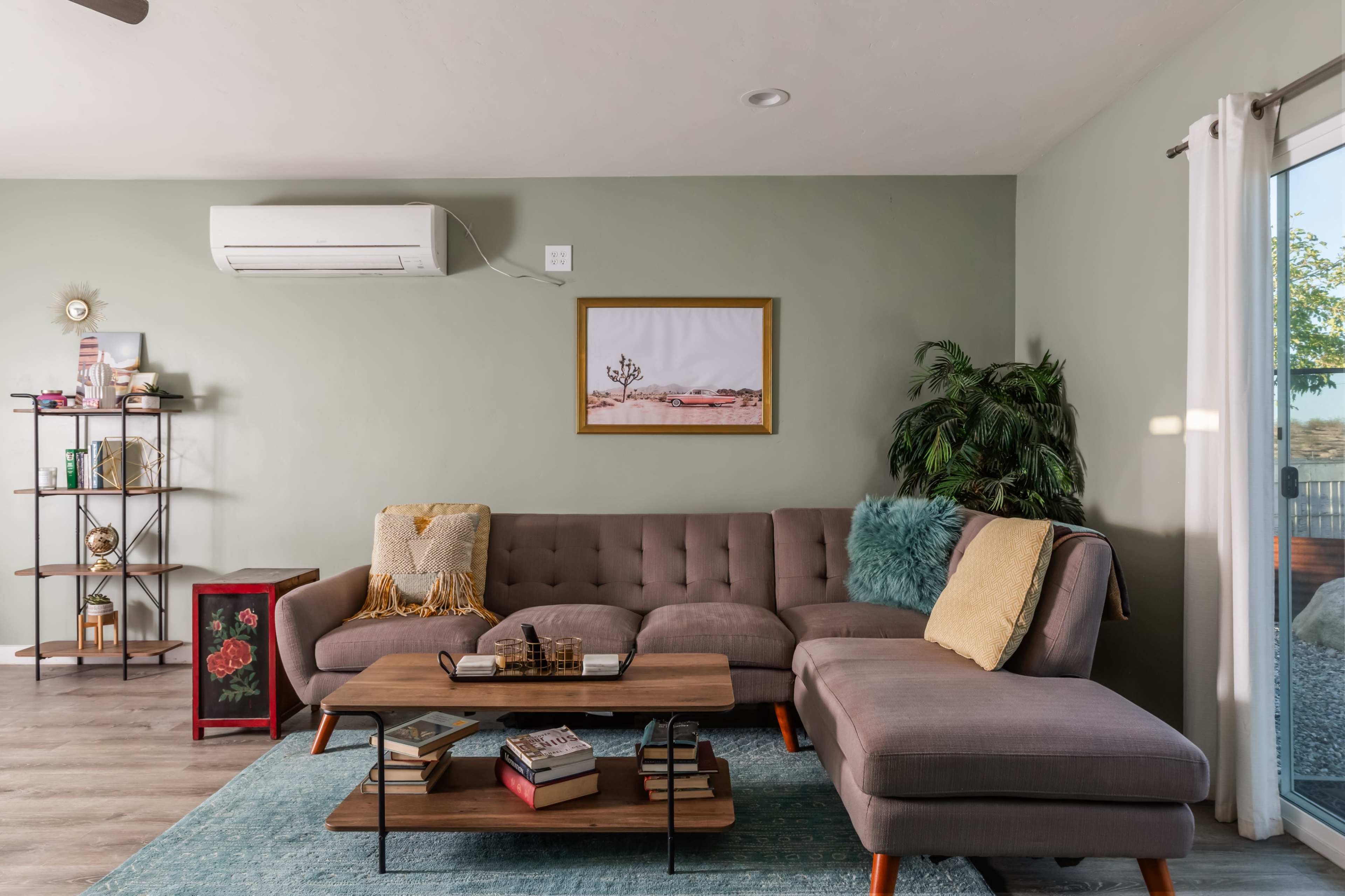The image shows a cozy living room with a gray sectional sofa, a wooden coffee table stacked with books, and a decorative plant beside a wall-mounted air conditioner.