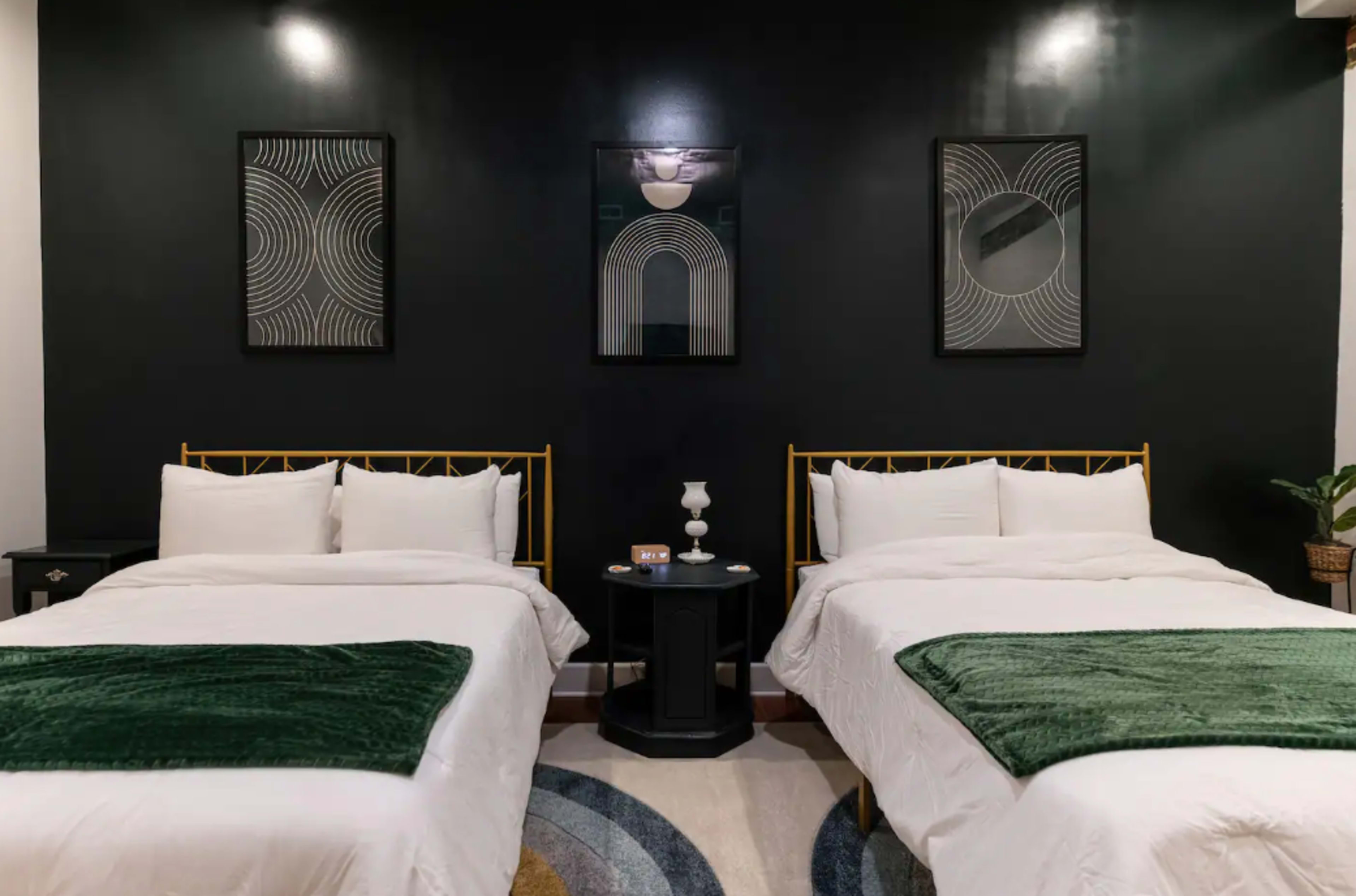 The image shows a room with two beds, each dressed in white linens and topped with green throws, flanking a small round table with decorative items against a dark wall adorned with abstract art.