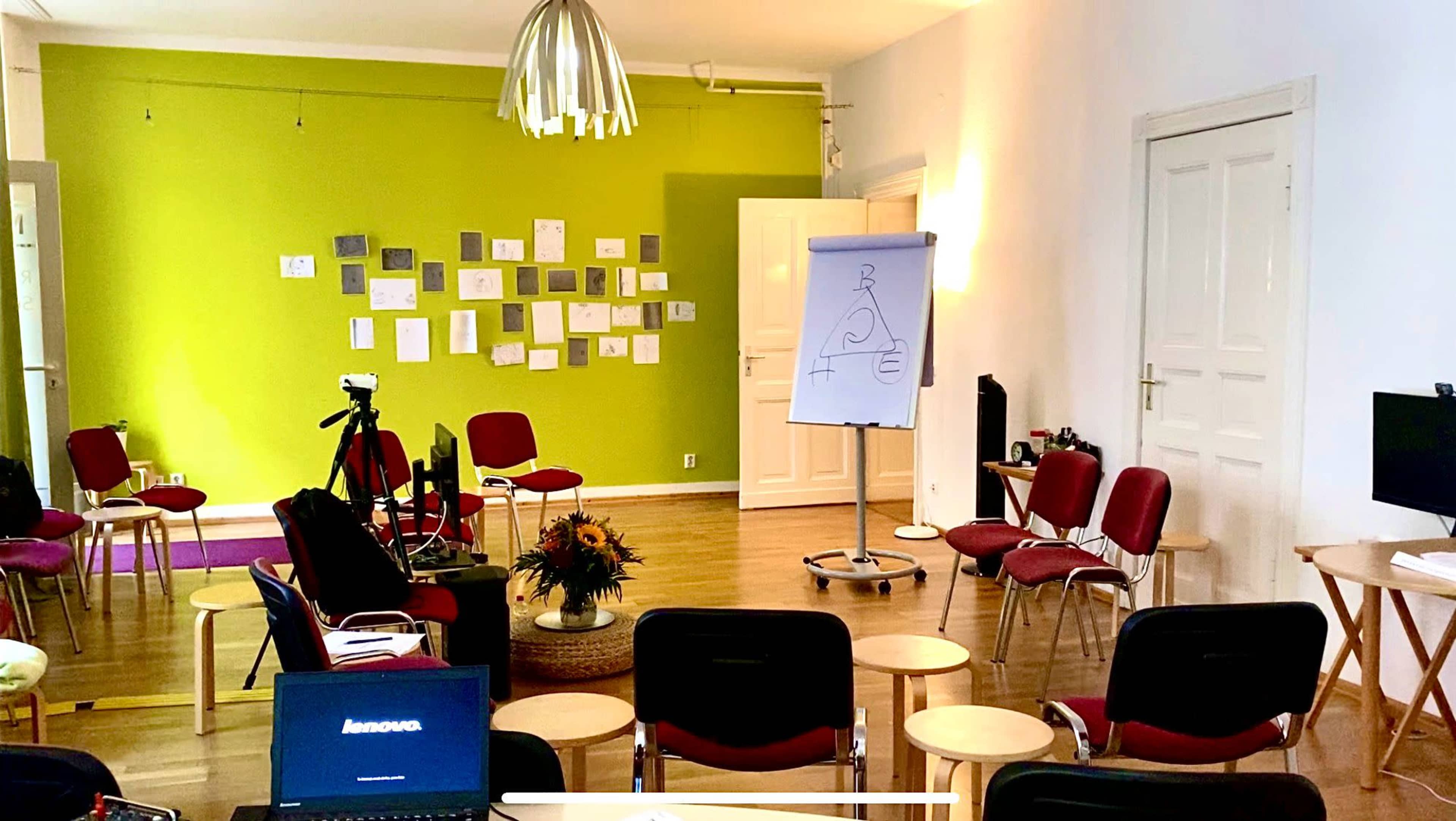 The image shows a brightly lit meeting room with green walls, several chairs arranged in a semi-circle, a white flip chart displaying a diagram, and various papers mounted on the wall.