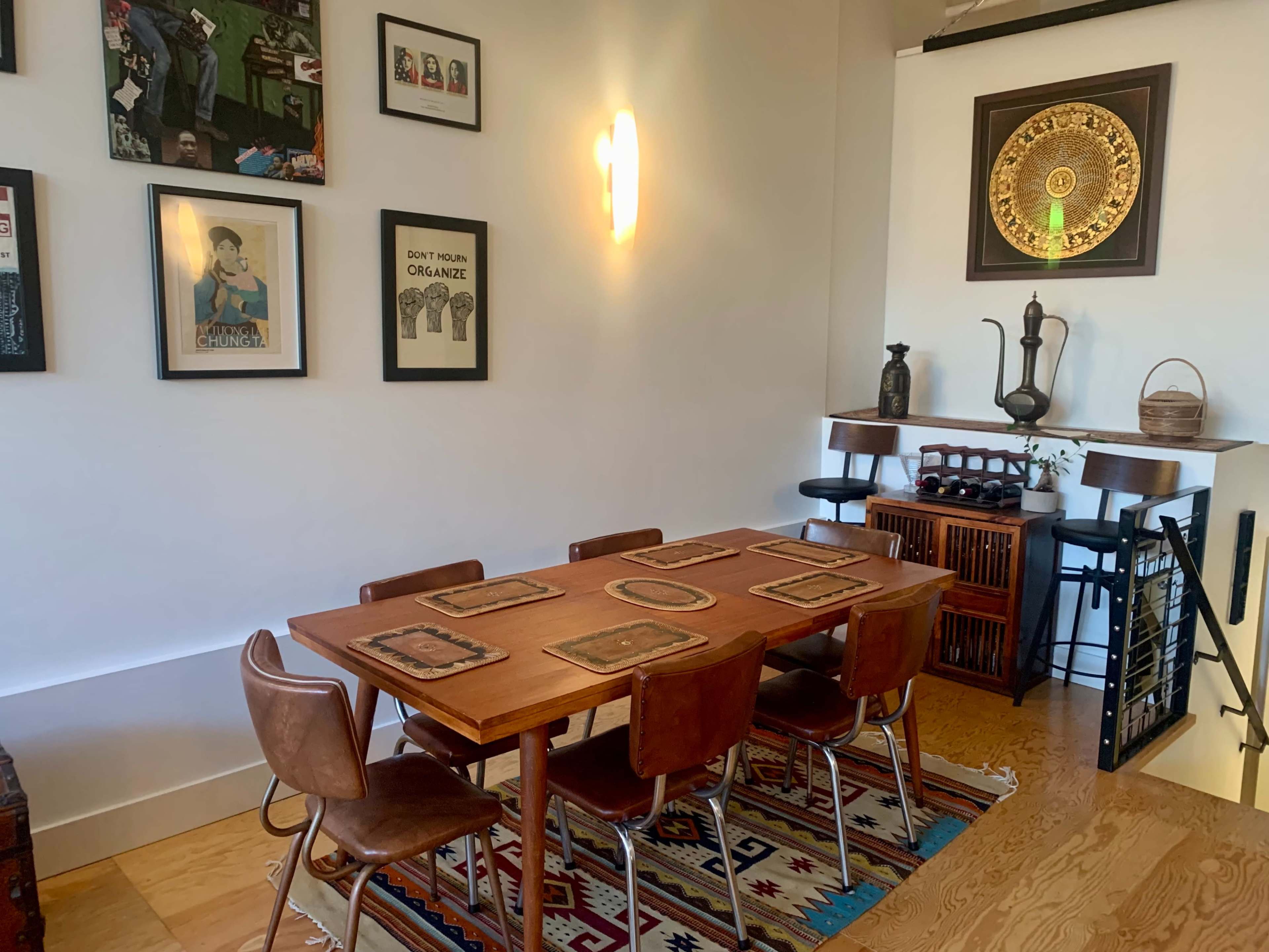 A wooden dining table with chairs is set in a brightly lit room adorned with framed artwork and decorative items on a shelf.