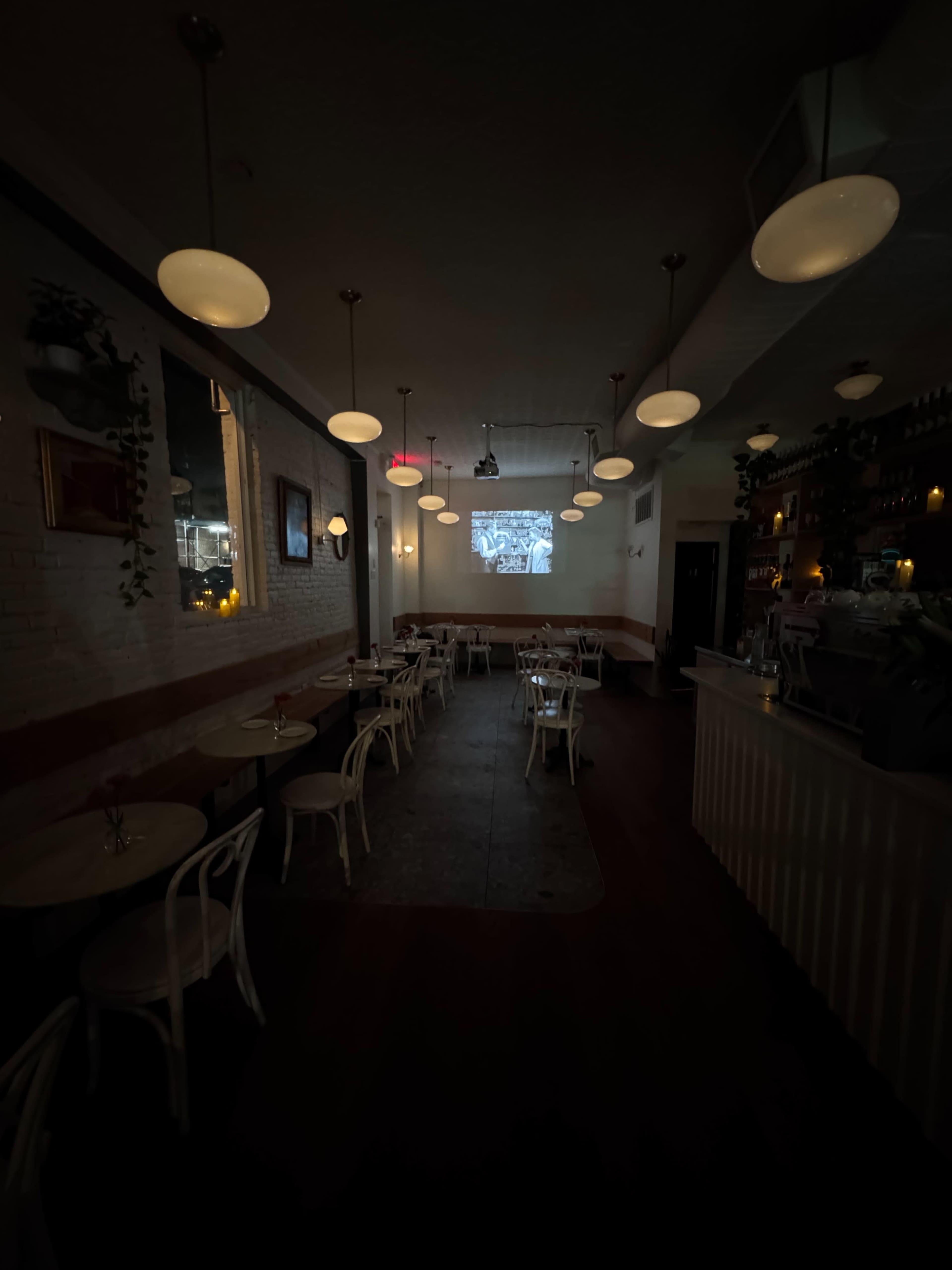 The image shows a dimly lit restaurant interior featuring white chairs and tables arranged in rows, with a projection screen displaying an image at the far end.