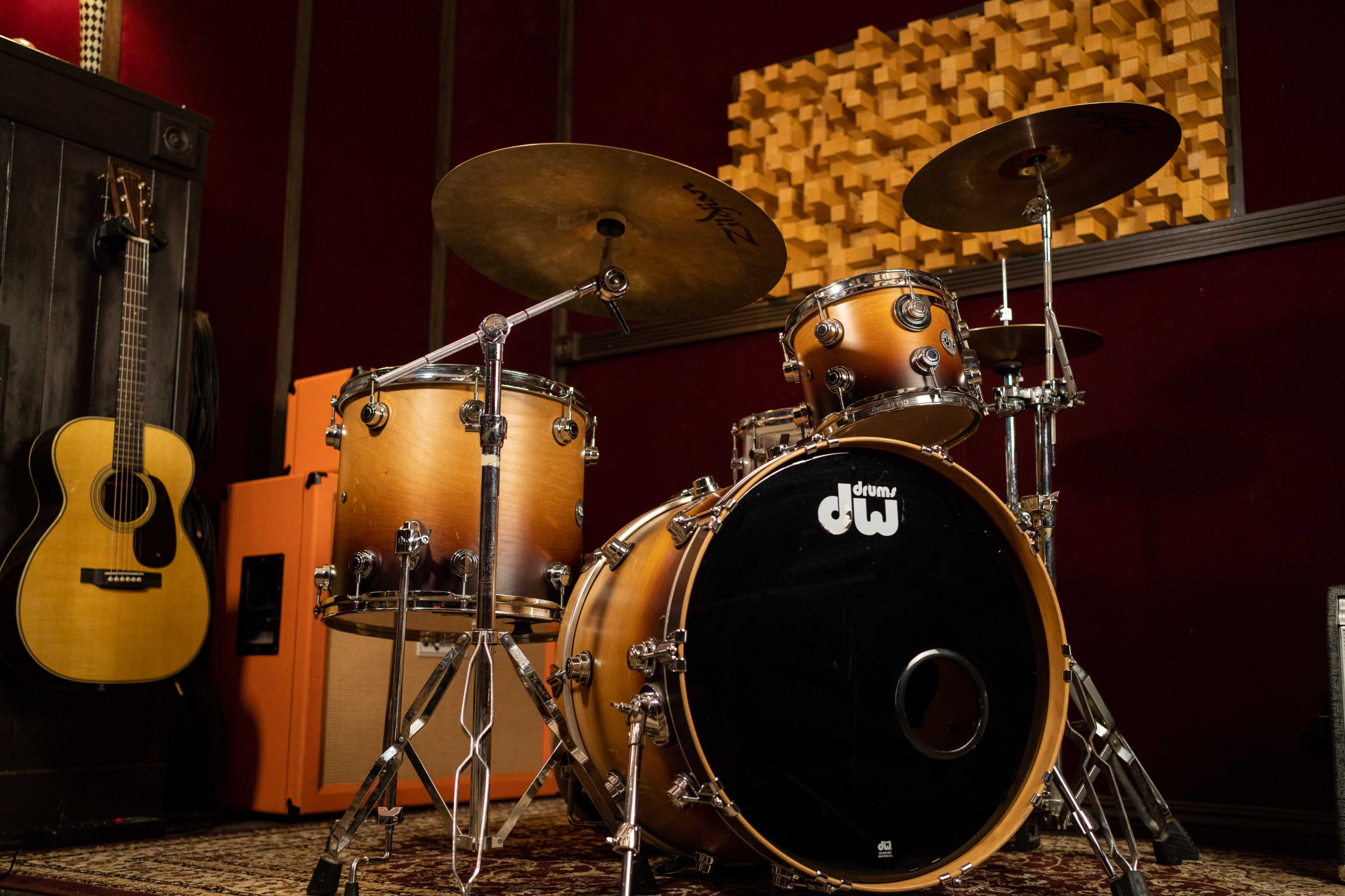 A drum set with a black drumhead and wooden finish, positioned in a music studio alongside an acoustic guitar and orange amplifiers.