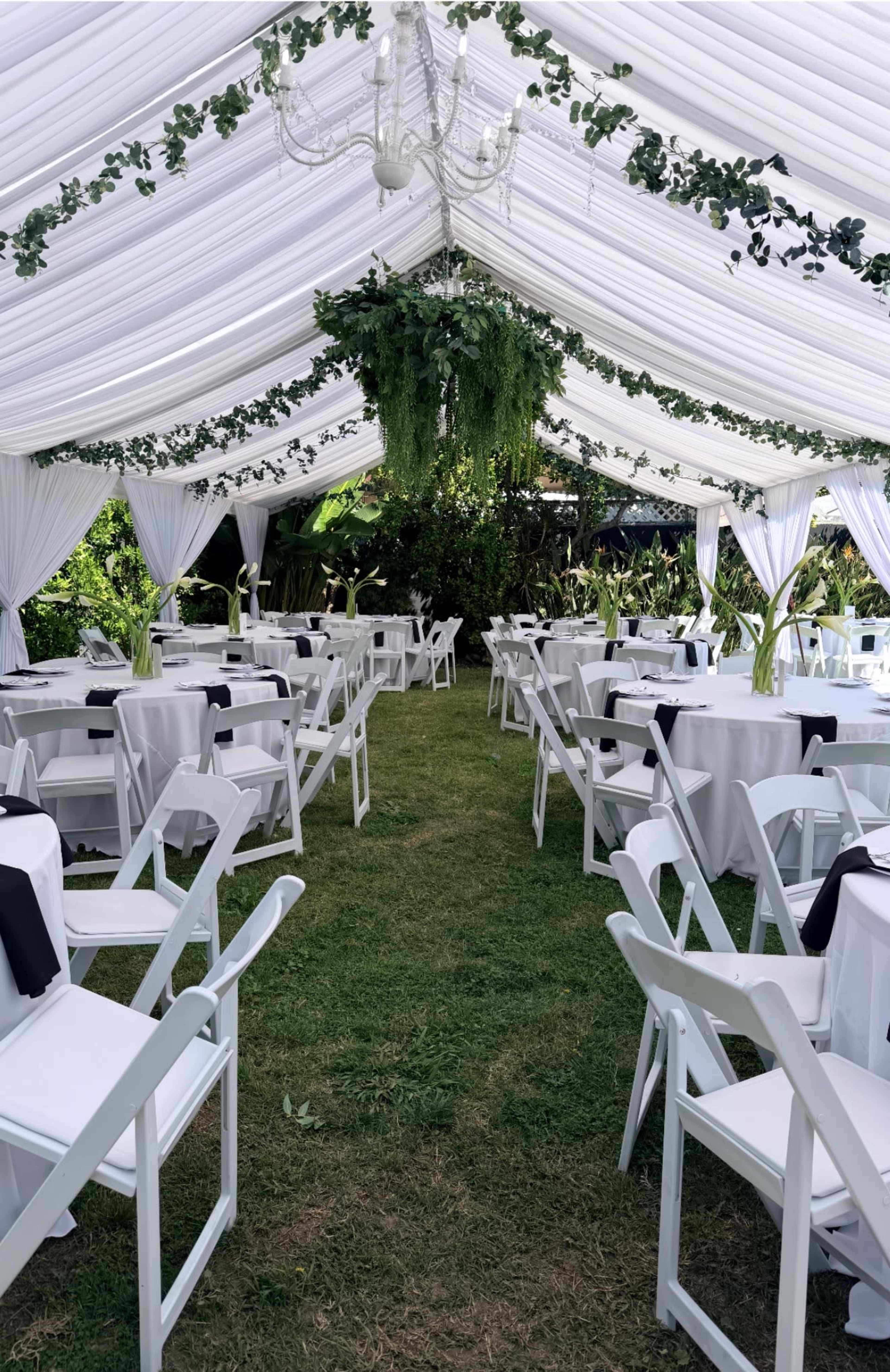 A white tent with decorated seating arranged in rows on green grass, featuring hanging greenery and a chandelier.