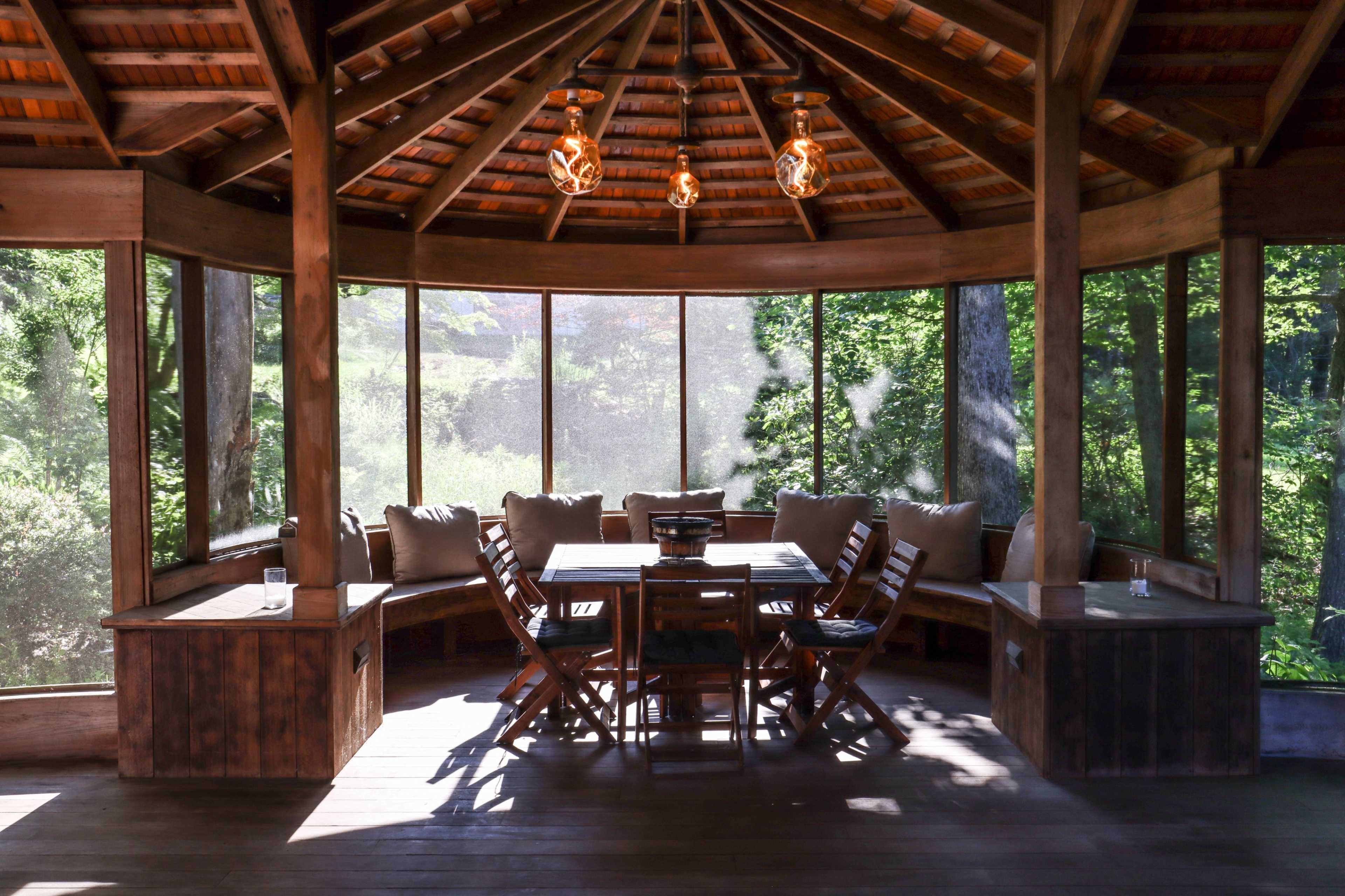 A wooden gazebo with large glass windows, furnished with a dining table and chairs surrounded by greenery.