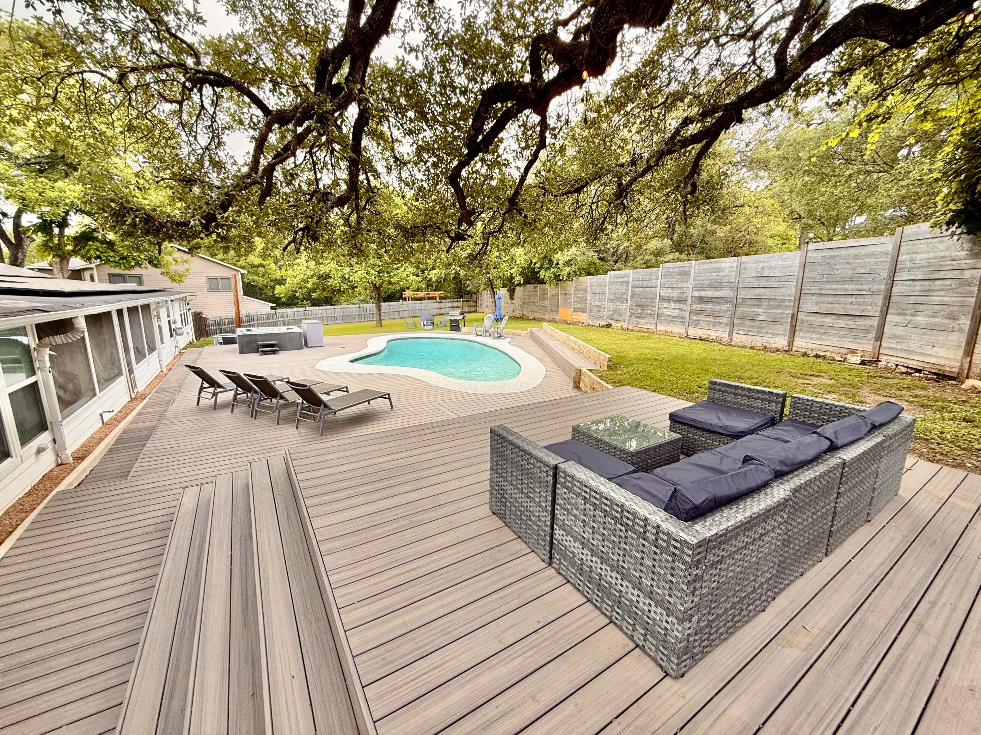 A spacious deck surrounds a swimming pool, featuring outdoor seating and lounge chairs, with a backdrop of greenery and wooden fencing.