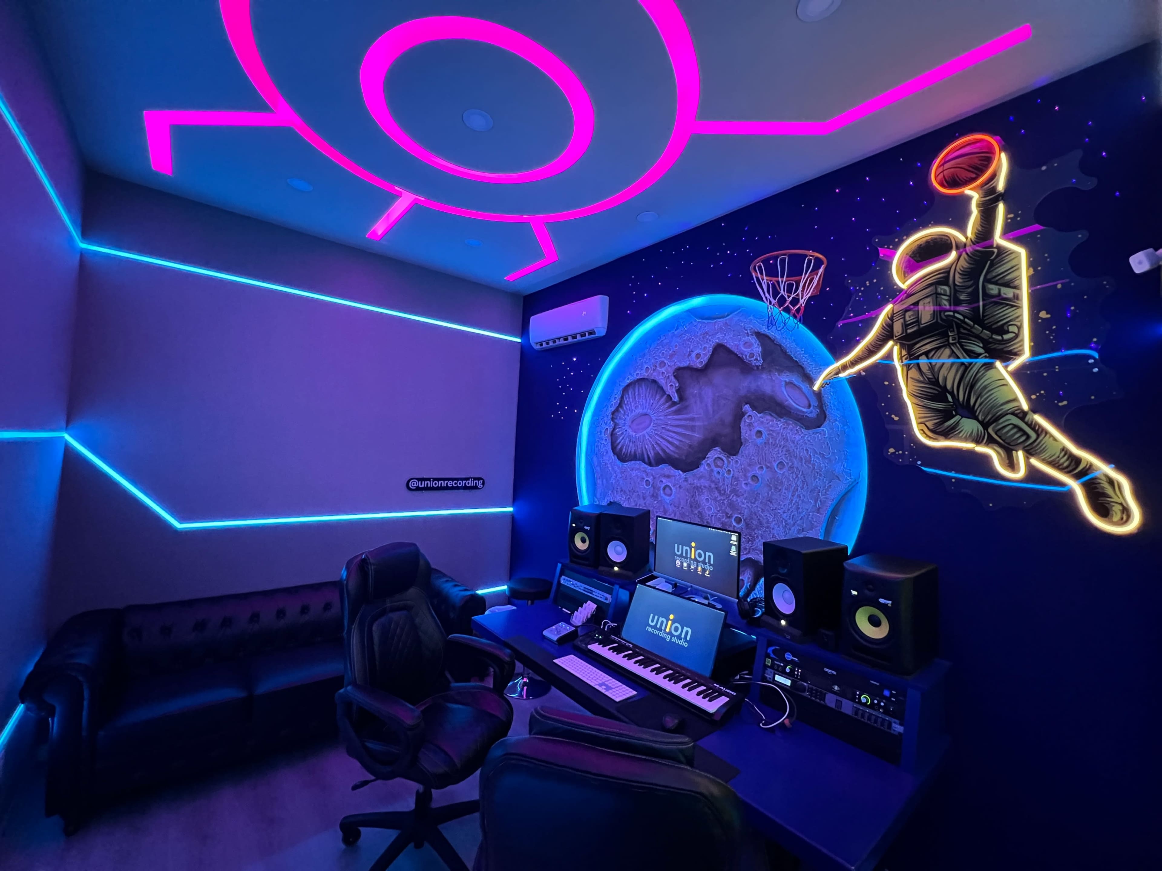 The image shows a modern music studio with neon blue and pink lights, a black sofa, a desk with audio equipment, and a mural of a basketball player against a space-themed backdrop.