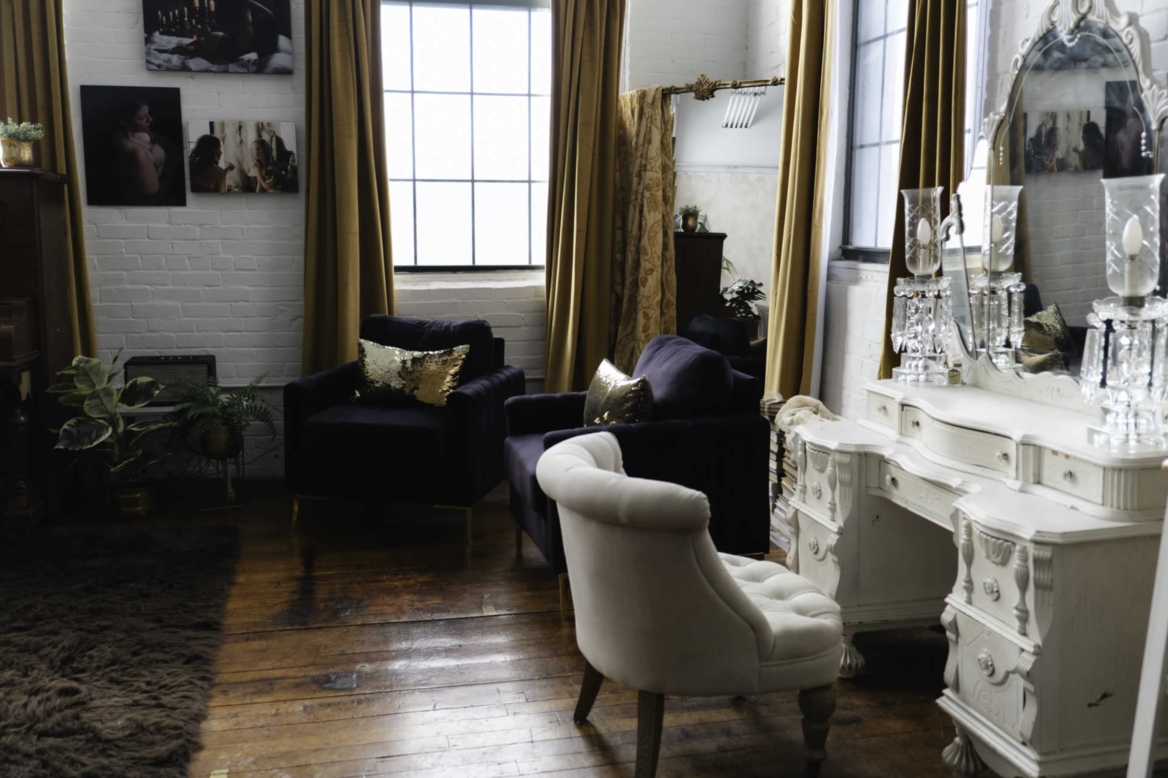 The image shows a styled living room with two dark blue armchairs, a white vanity, and large windows draped with golden curtains.