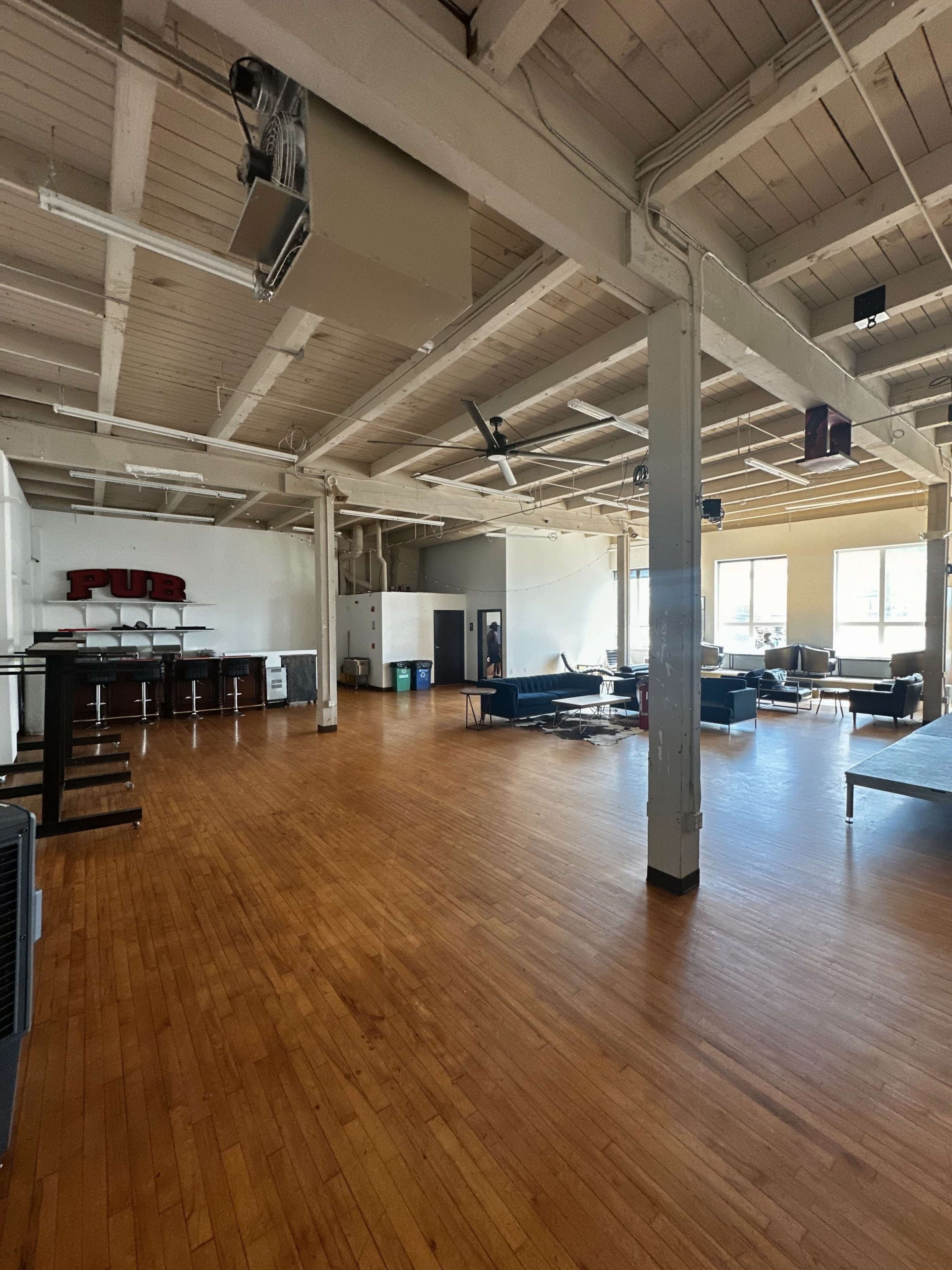 The image shows an empty, spacious room with wooden floors, large windows, and various pieces of furniture, including sofas and tables.