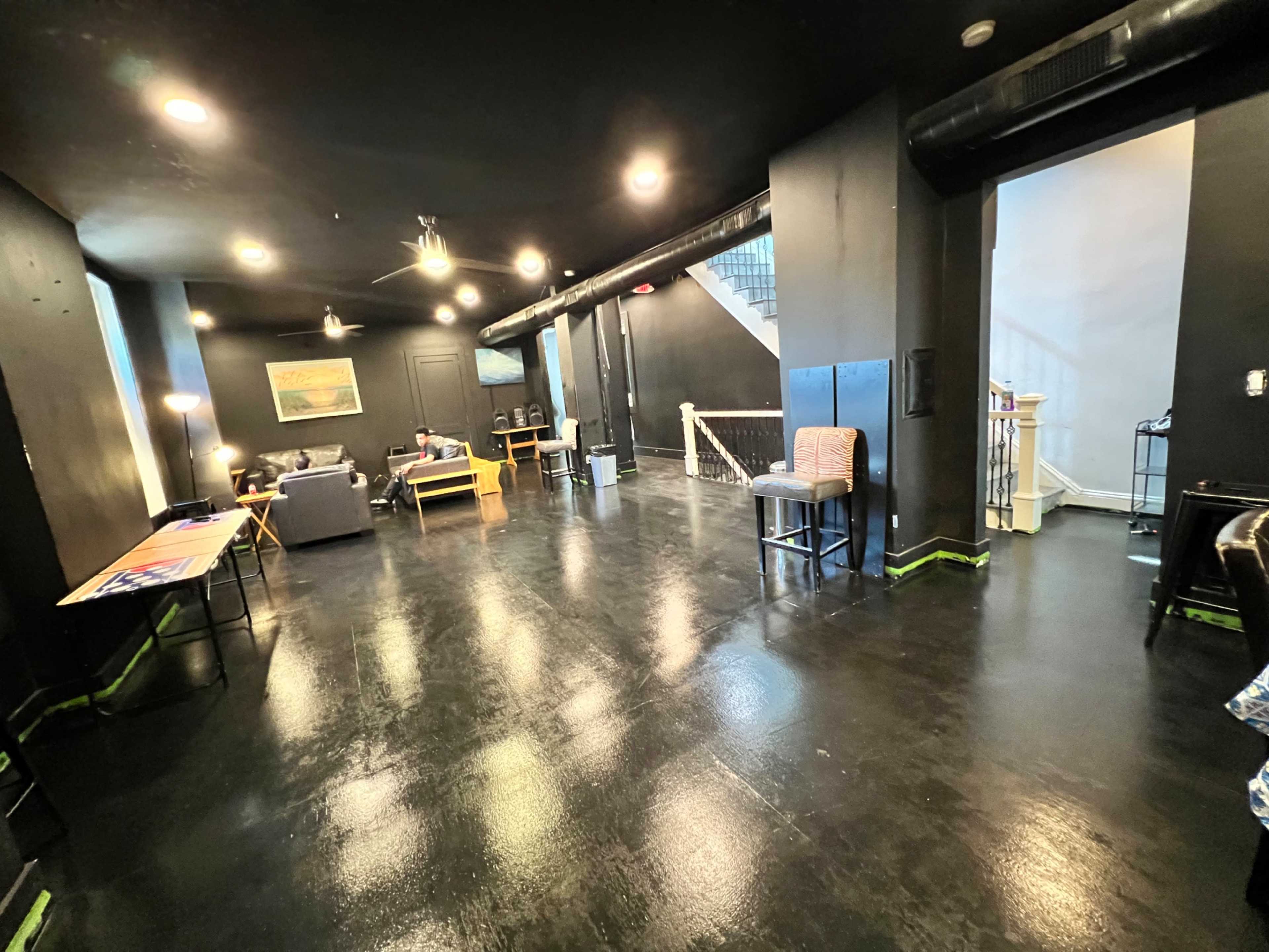 A spacious, dimly lit room with black walls and polished floors, featuring scattered furniture such as sofas and tables along with a staircase in the background.