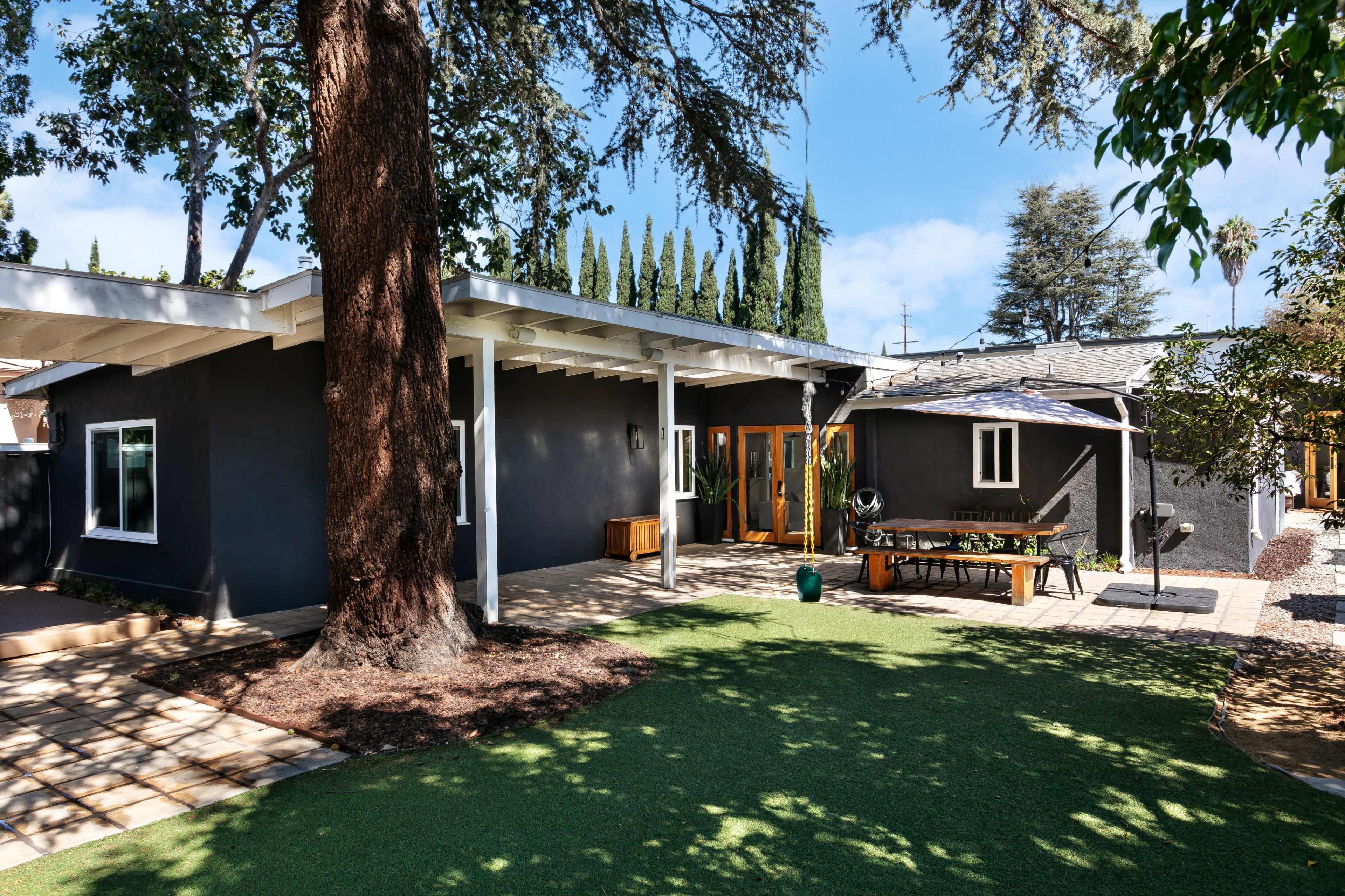 A modern single-story house with a black exterior, surrounded by greenery and a patio area featuring a table and benches.