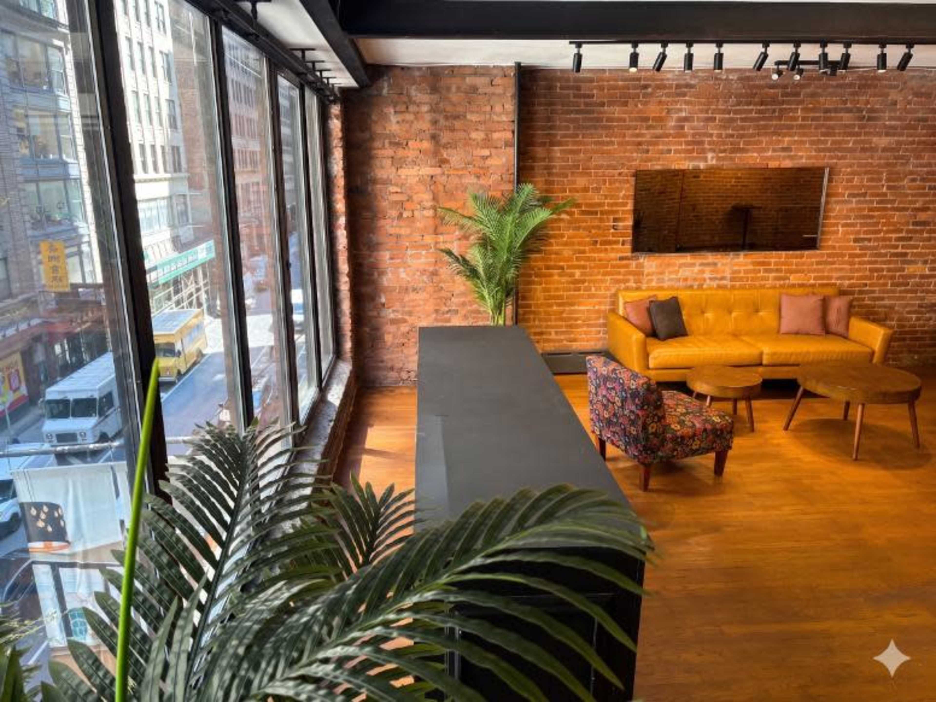 The image shows a modern interior space with large windows, exposed brick walls, a yellow sofa, a patterned chair, and a dark counter, alongside a potted palm plant.