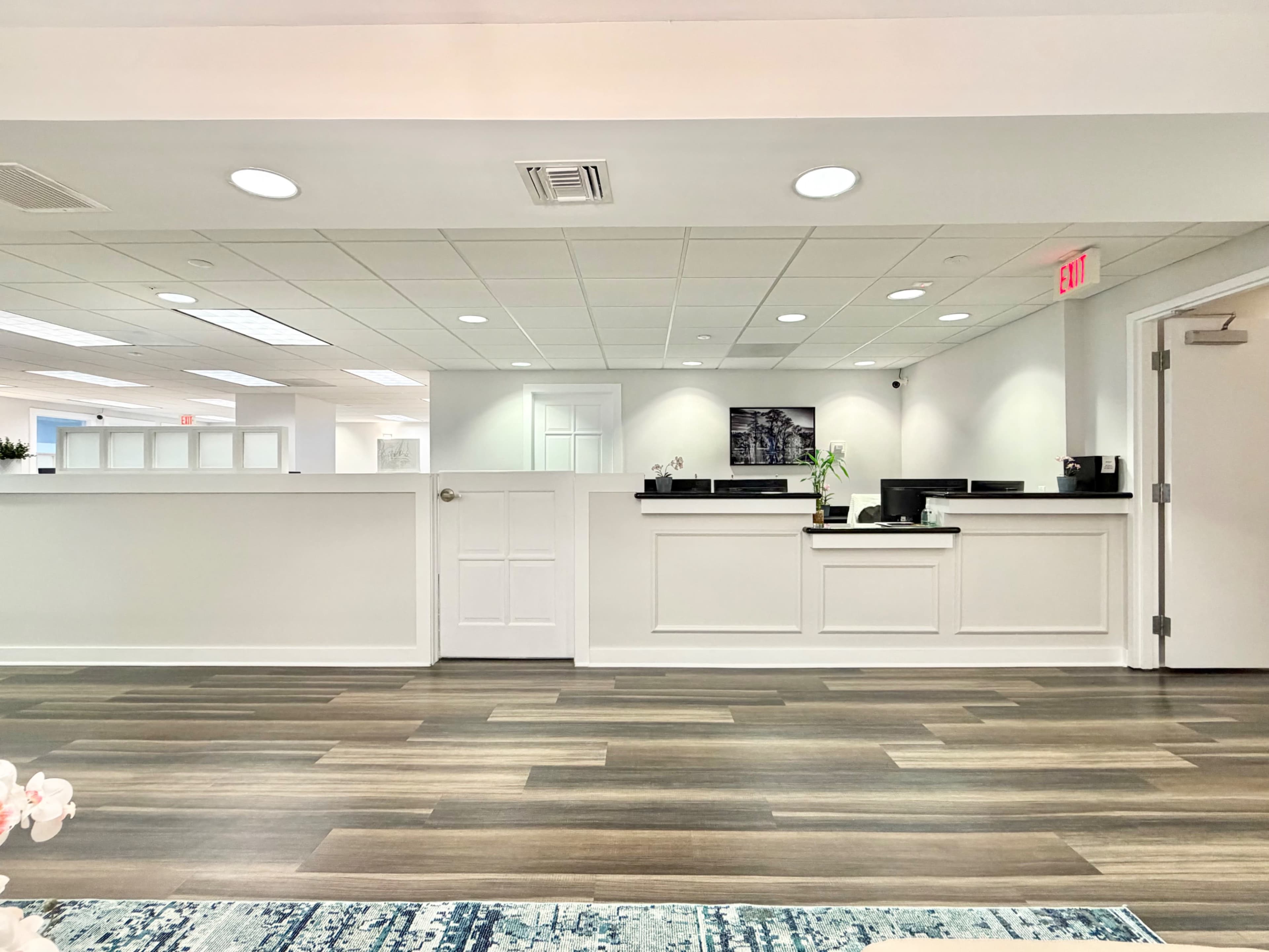 The image shows a modern office reception area with a white counter and dark wood flooring, featuring several computer stations and decorative plants.
