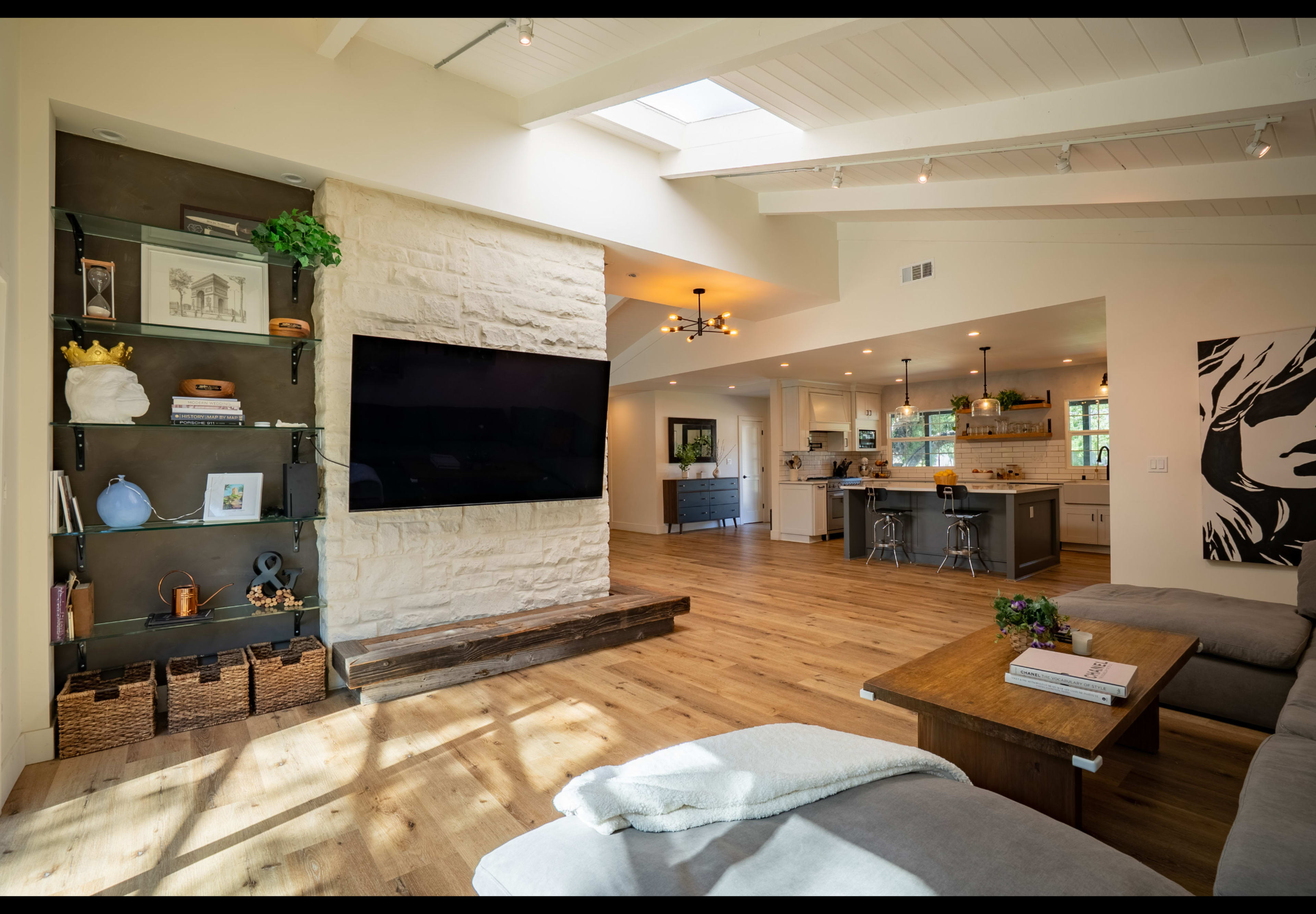 A bright living room features a large mounted TV, a stone accent wall, and open access to a kitchen with wooden floors and modern furnishings.