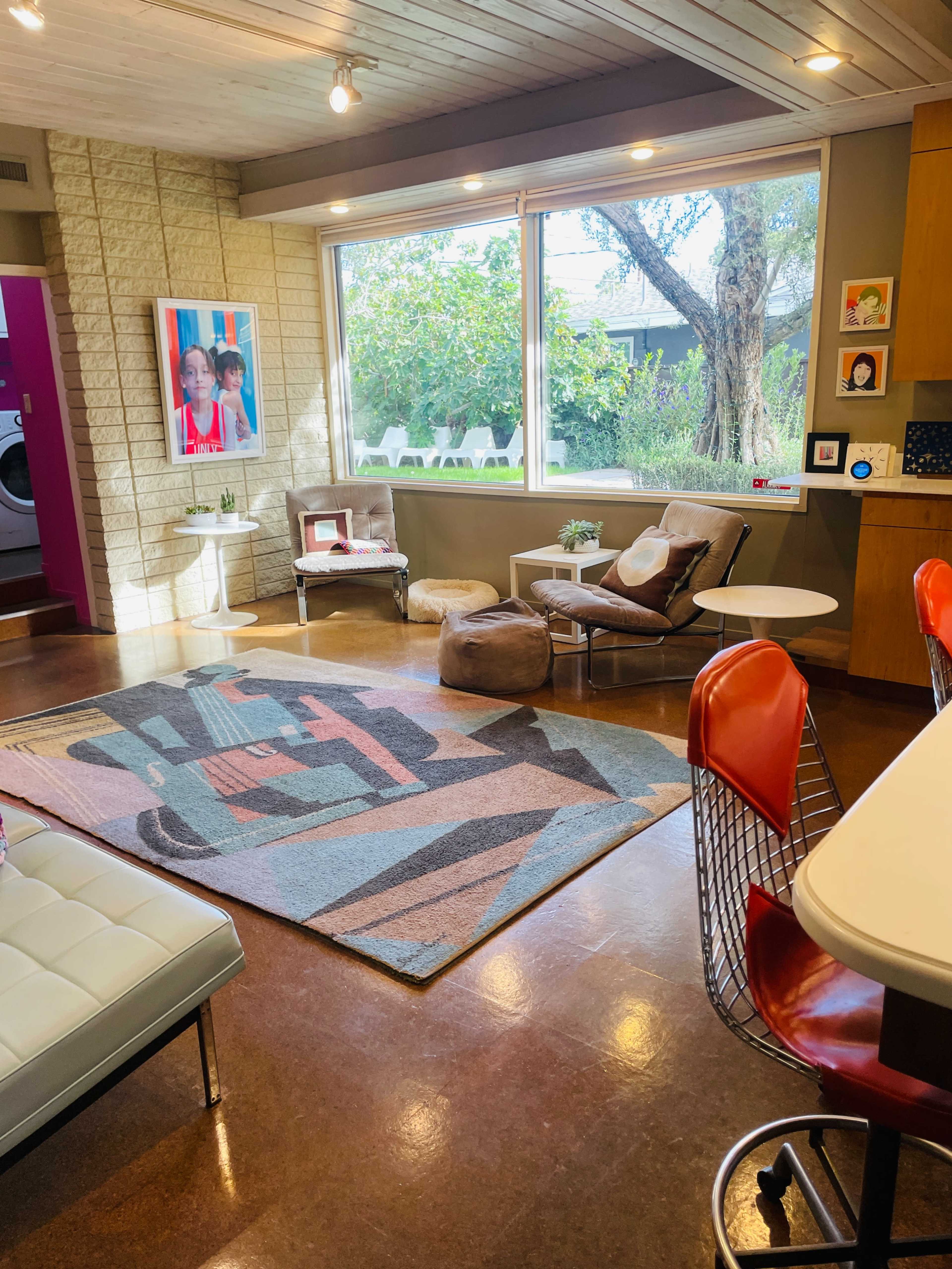 The image shows a modern living room with a large window, a colorful area rug, minimalist furniture, and decorative art on the walls.