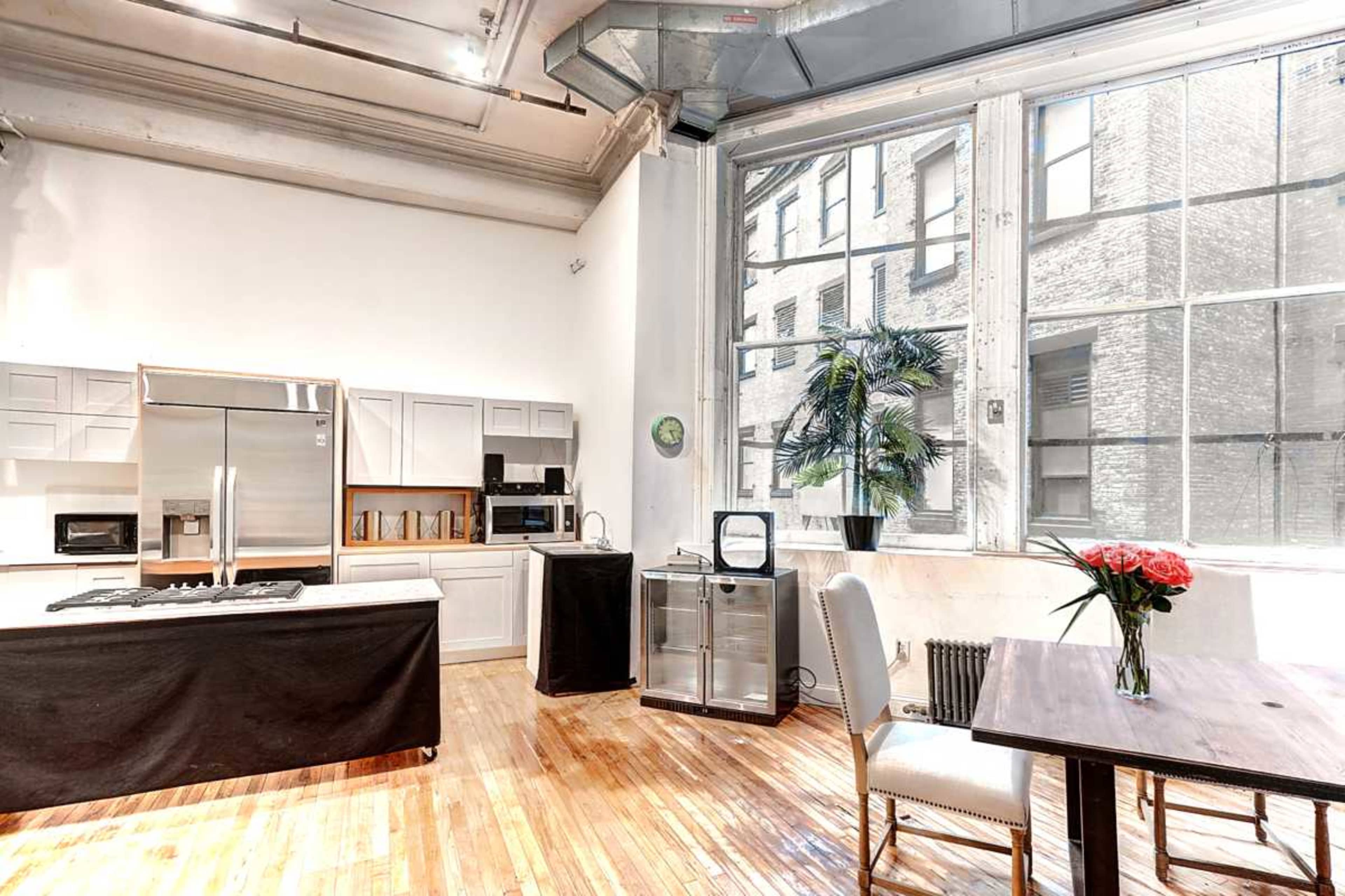 The image shows a bright, open living space with a kitchen area, large windows, and a dining table with flowers.