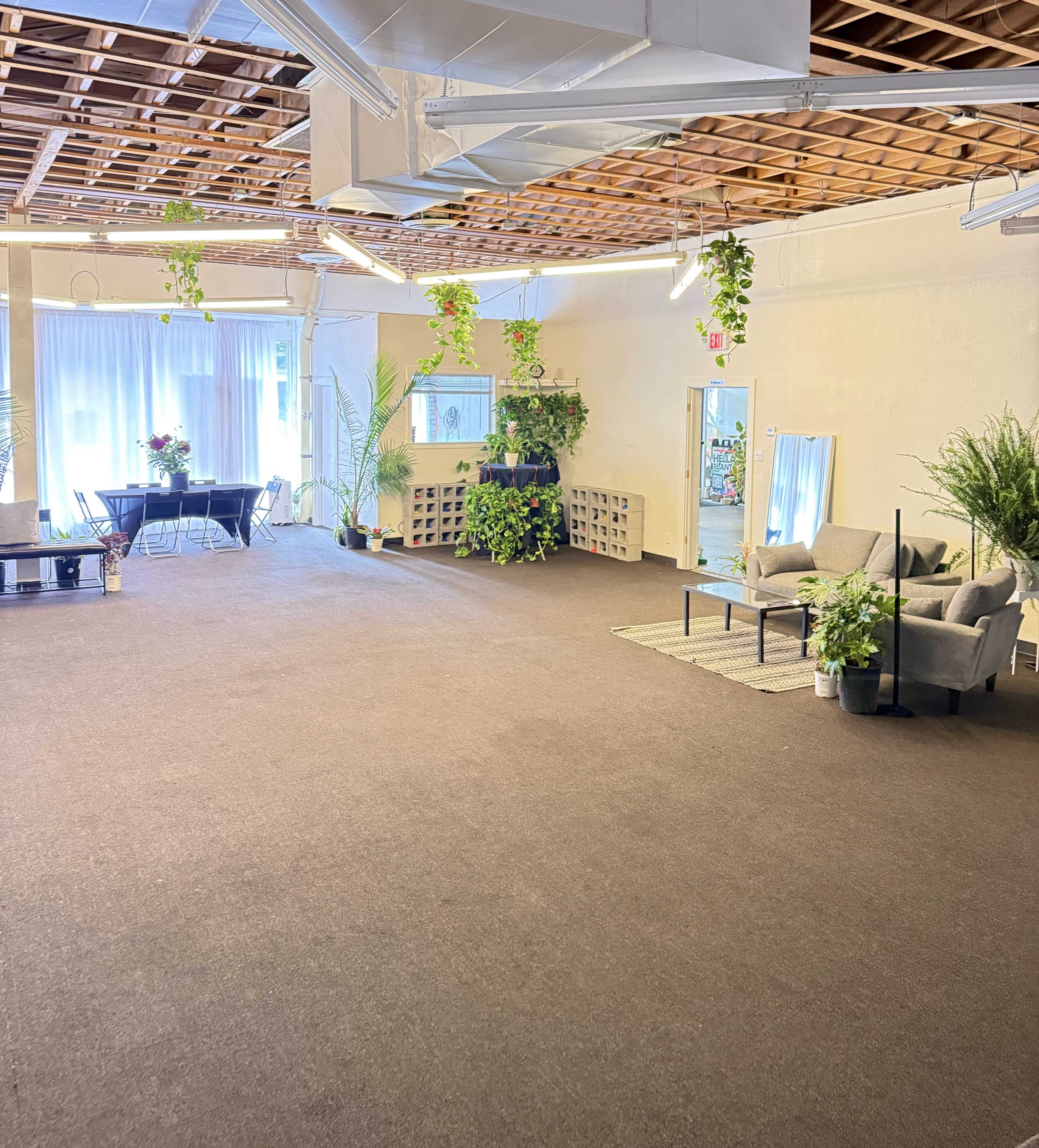 The image shows a spacious, well-lit room with plants, seating areas, and a table set for an event.