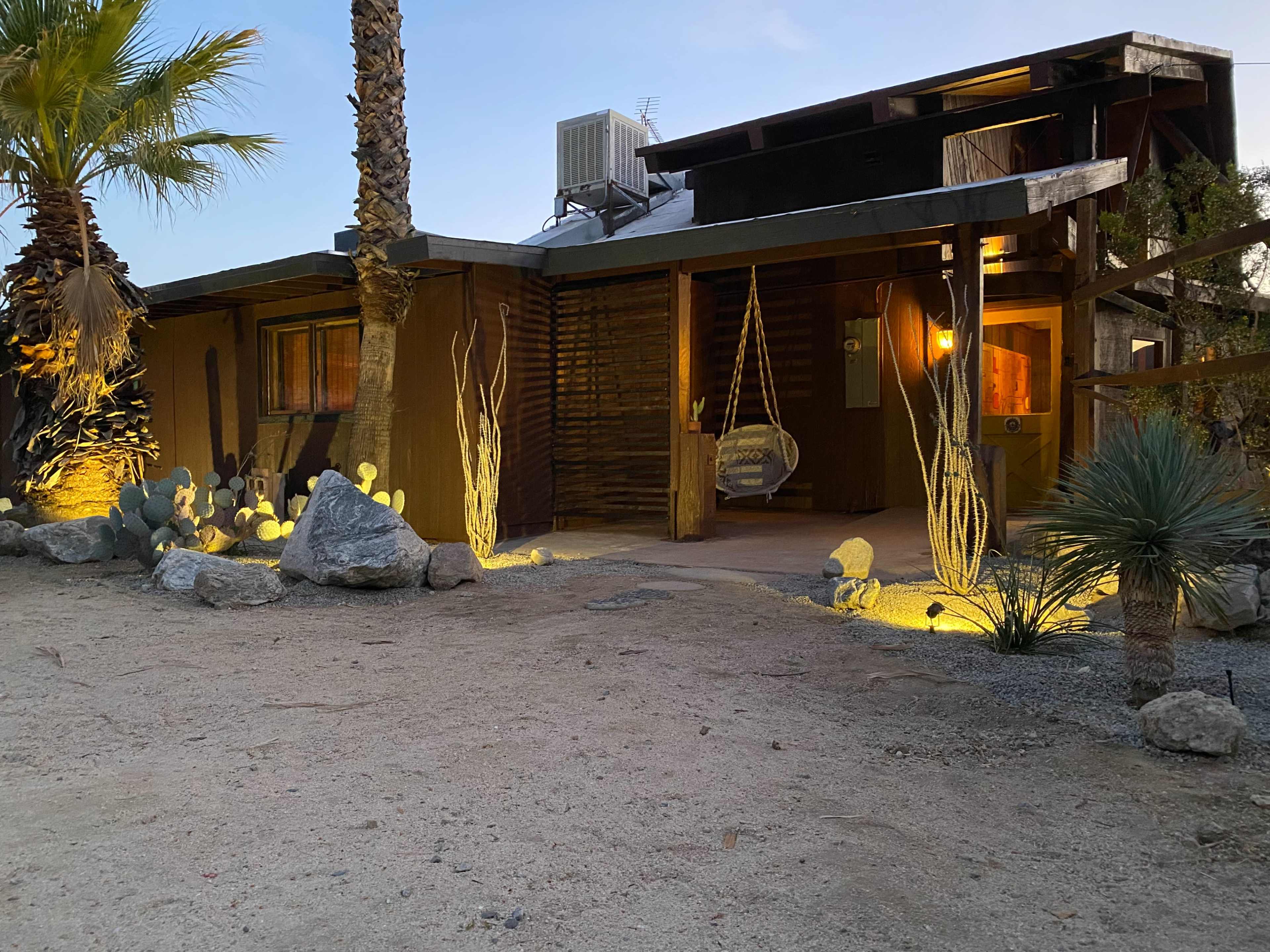 A rustic wooden house with a porch swing, surrounded by desert vegetation and illuminated by soft outdoor lighting.