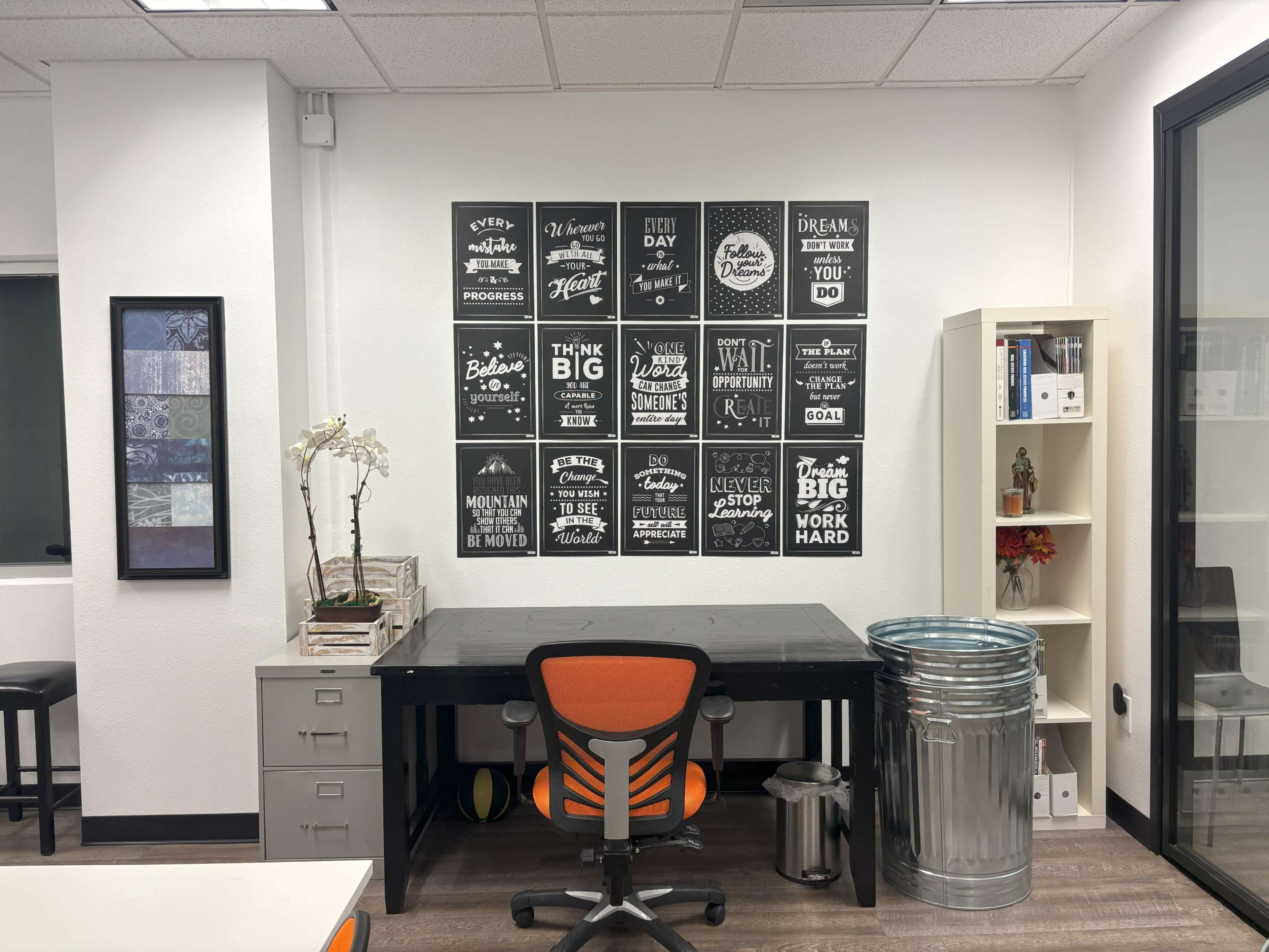 The image shows an office space with a black desk and an orange office chair, featuring motivational posters on the wall and storage cabinets beside the desk.