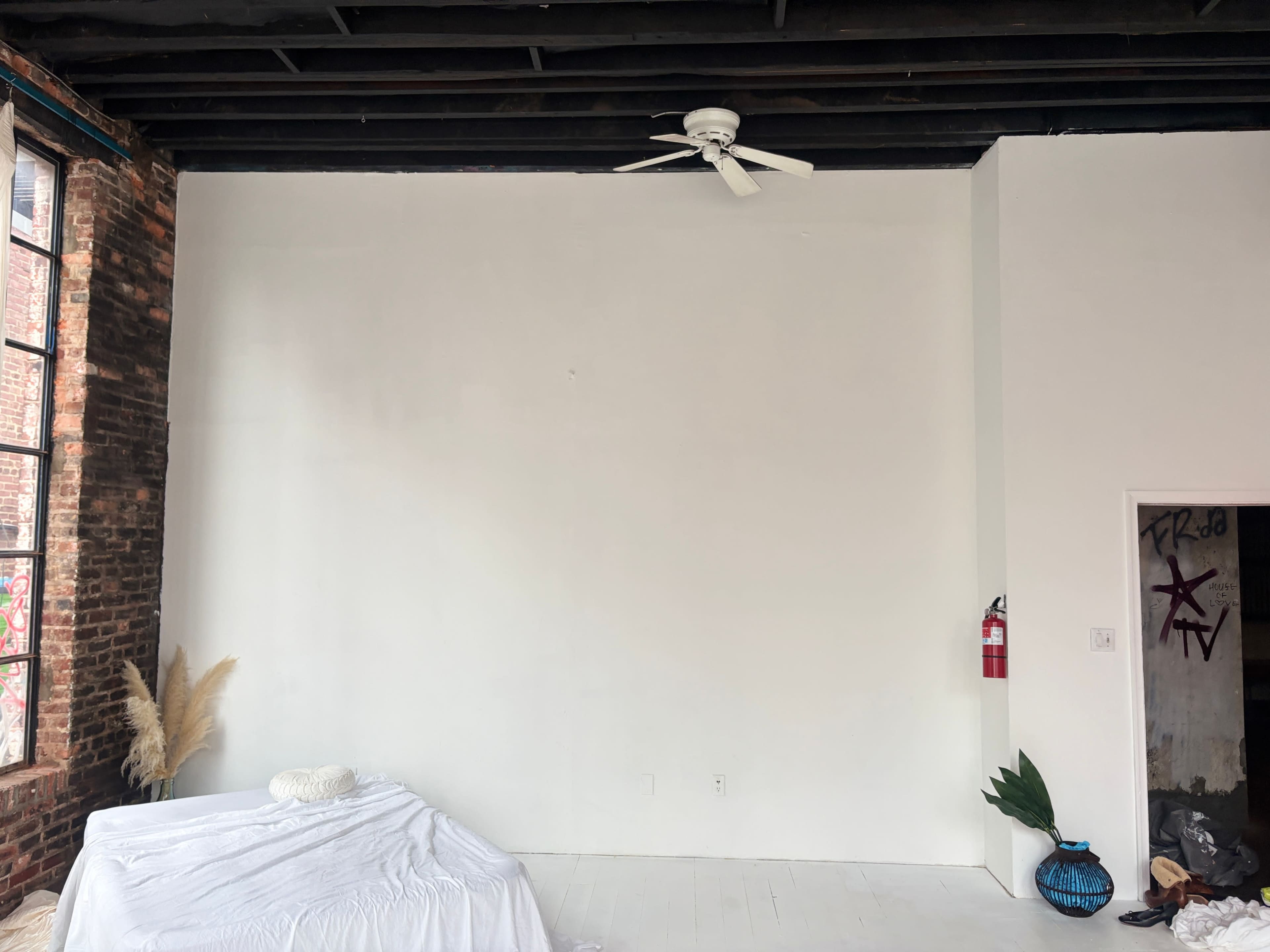 The image shows a spacious room with exposed brick walls, a white ceiling fan, a light-colored floor, a bed covered with a white sheet, and a door leading to another space.