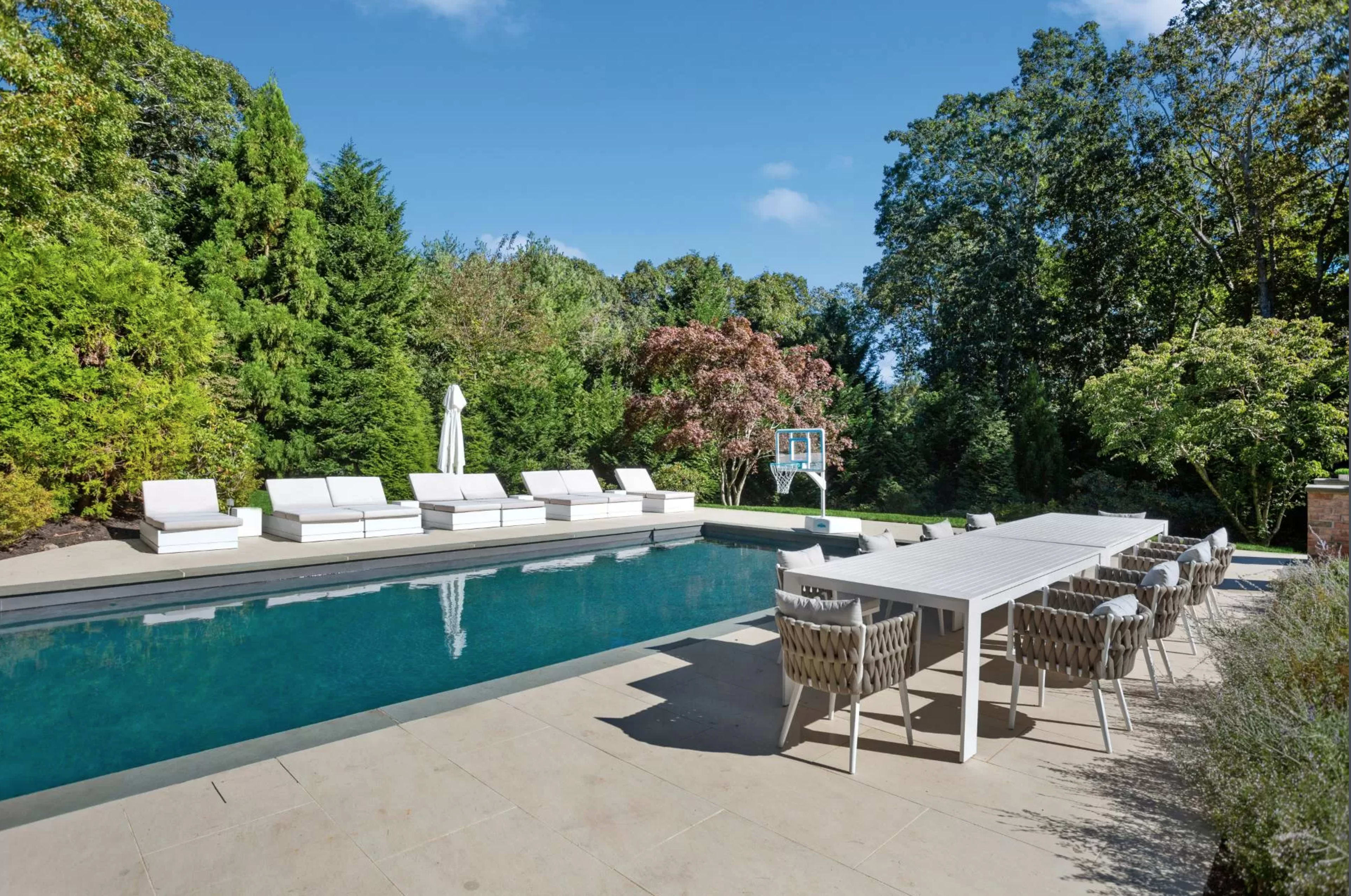 A modern outdoor area featuring a rectangular swimming pool surrounded by white lounge chairs and a long dining table, set against a backdrop of lush greenery.