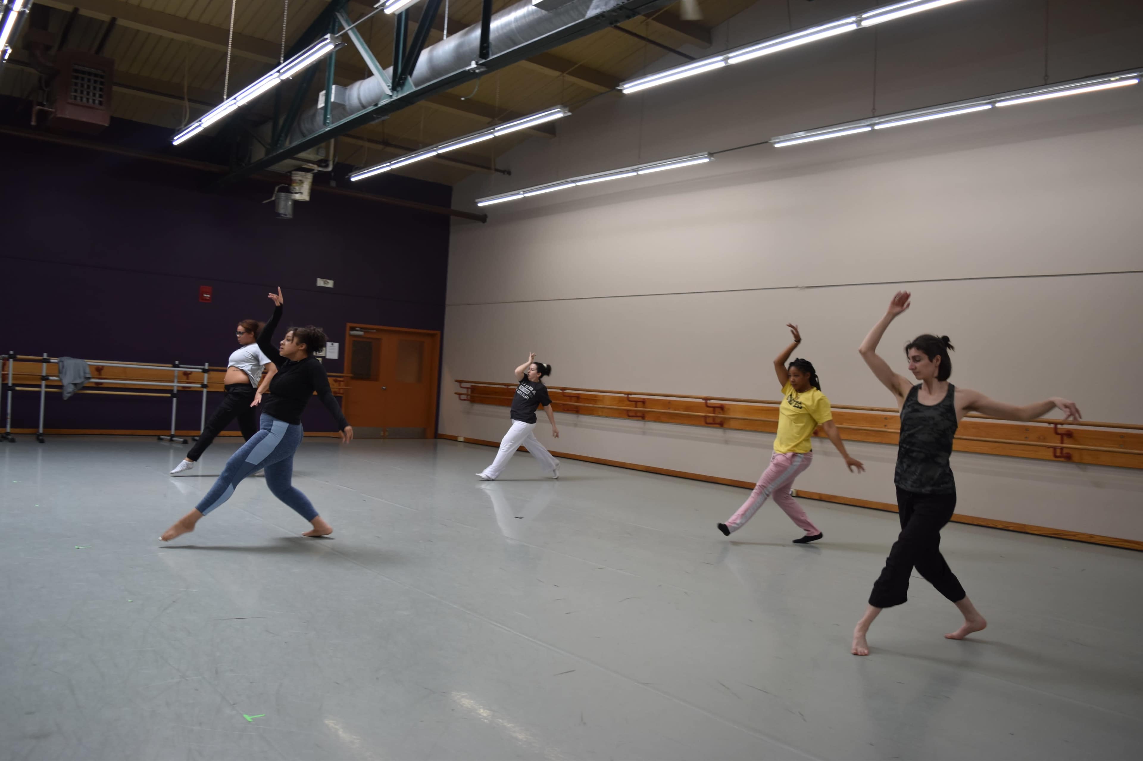 Five dancers are practicing in a well-lit studio with mirrored walls and wooden barre fixtures.