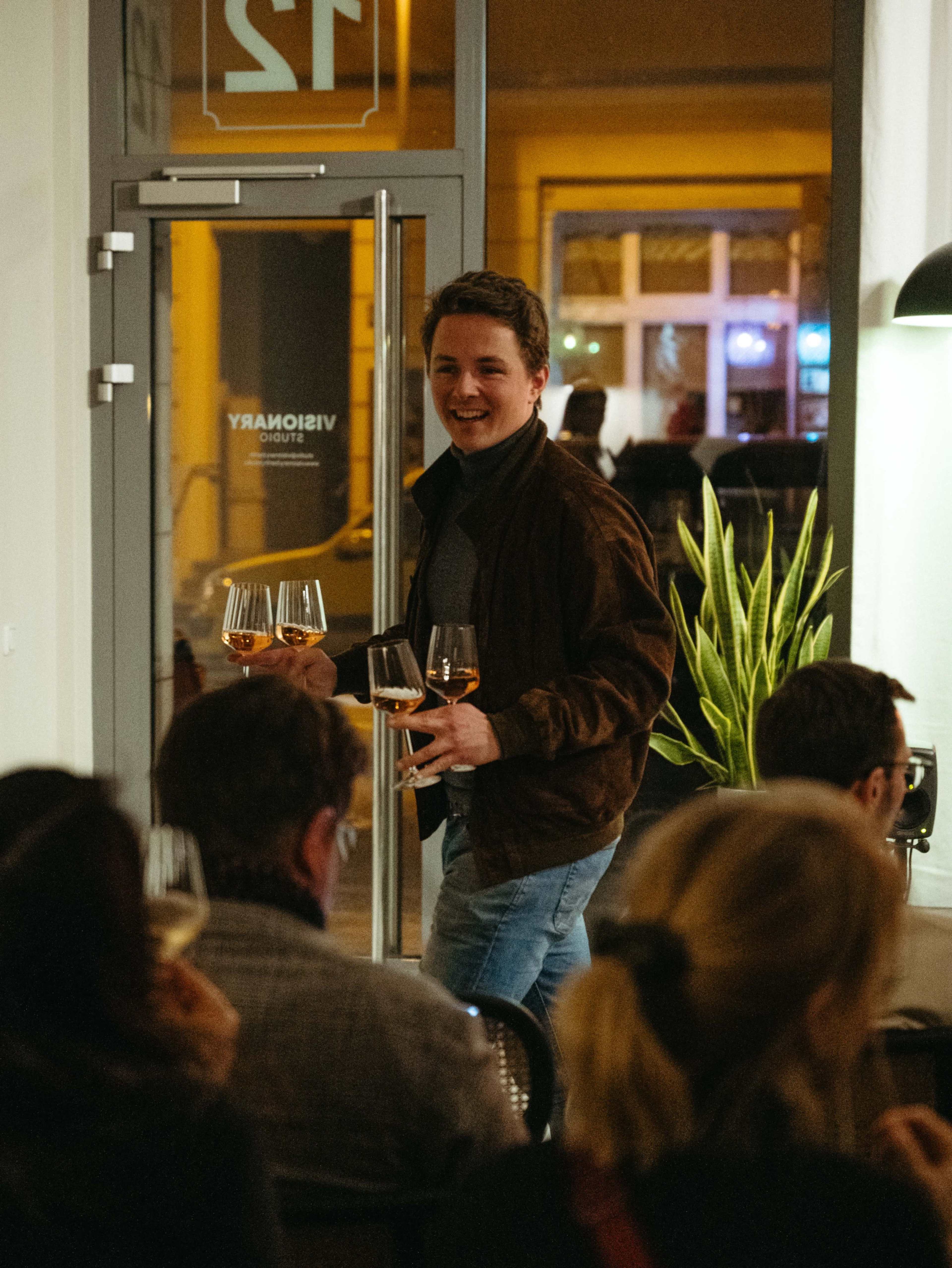 A man carries three glasses of drink while walking through a room filled with seated guests.