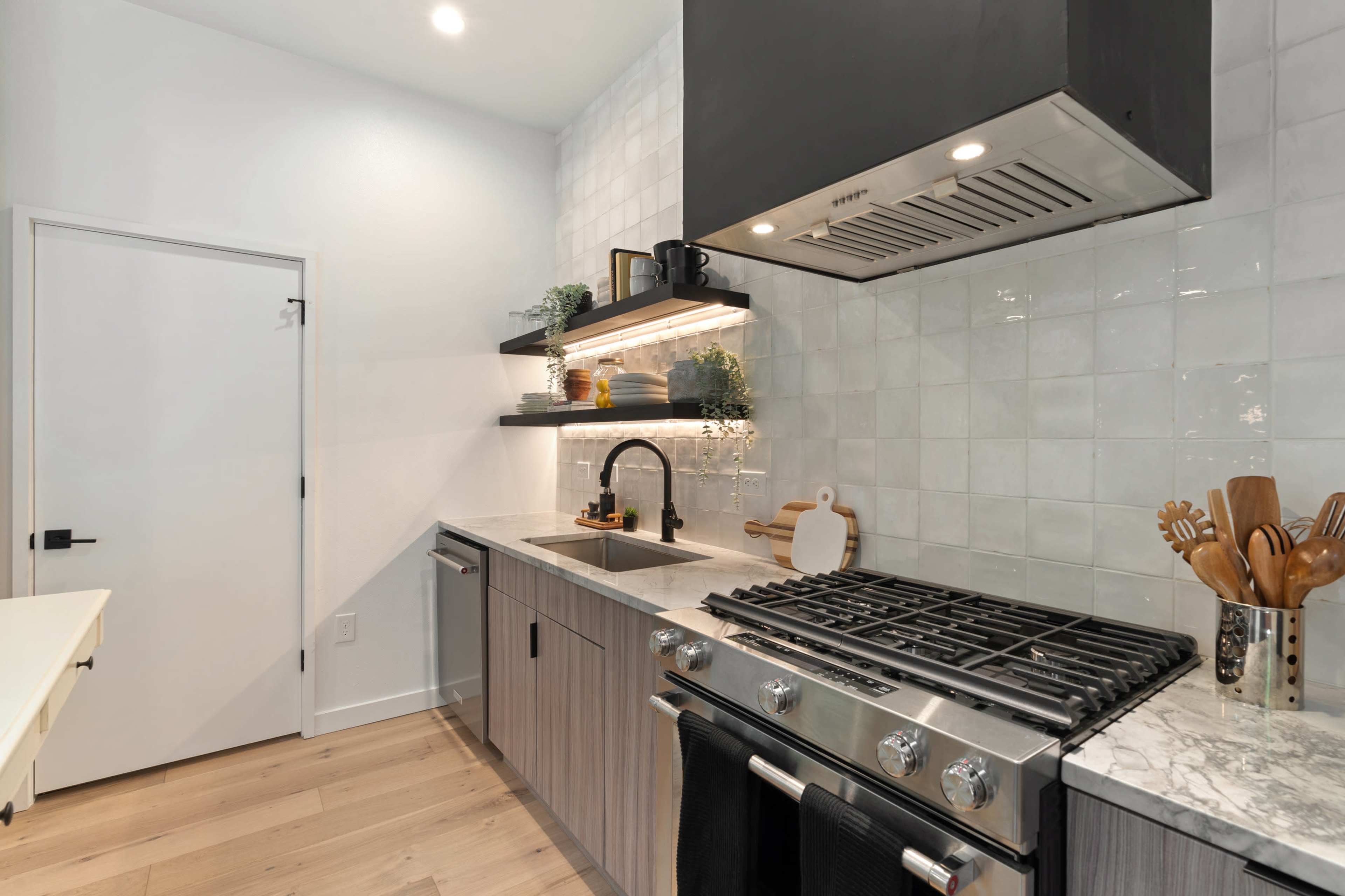 A modern kitchen with a stainless steel stove, a hood, wooden cabinets, and white tiled walls featuring open shelves with plants and kitchenware.