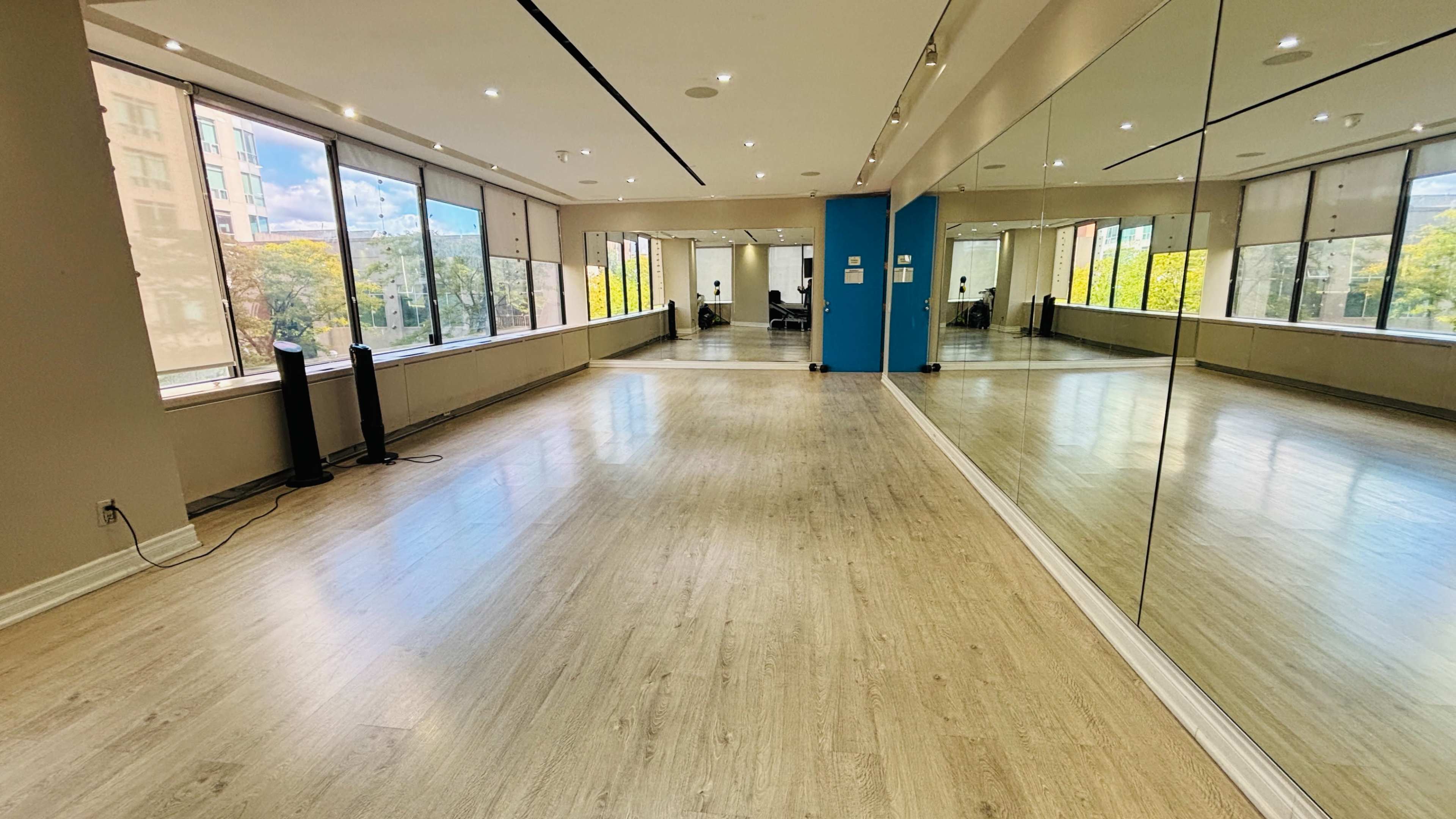 The image shows a spacious dance studio with large windows, a mirrored wall, and a light wooden floor.