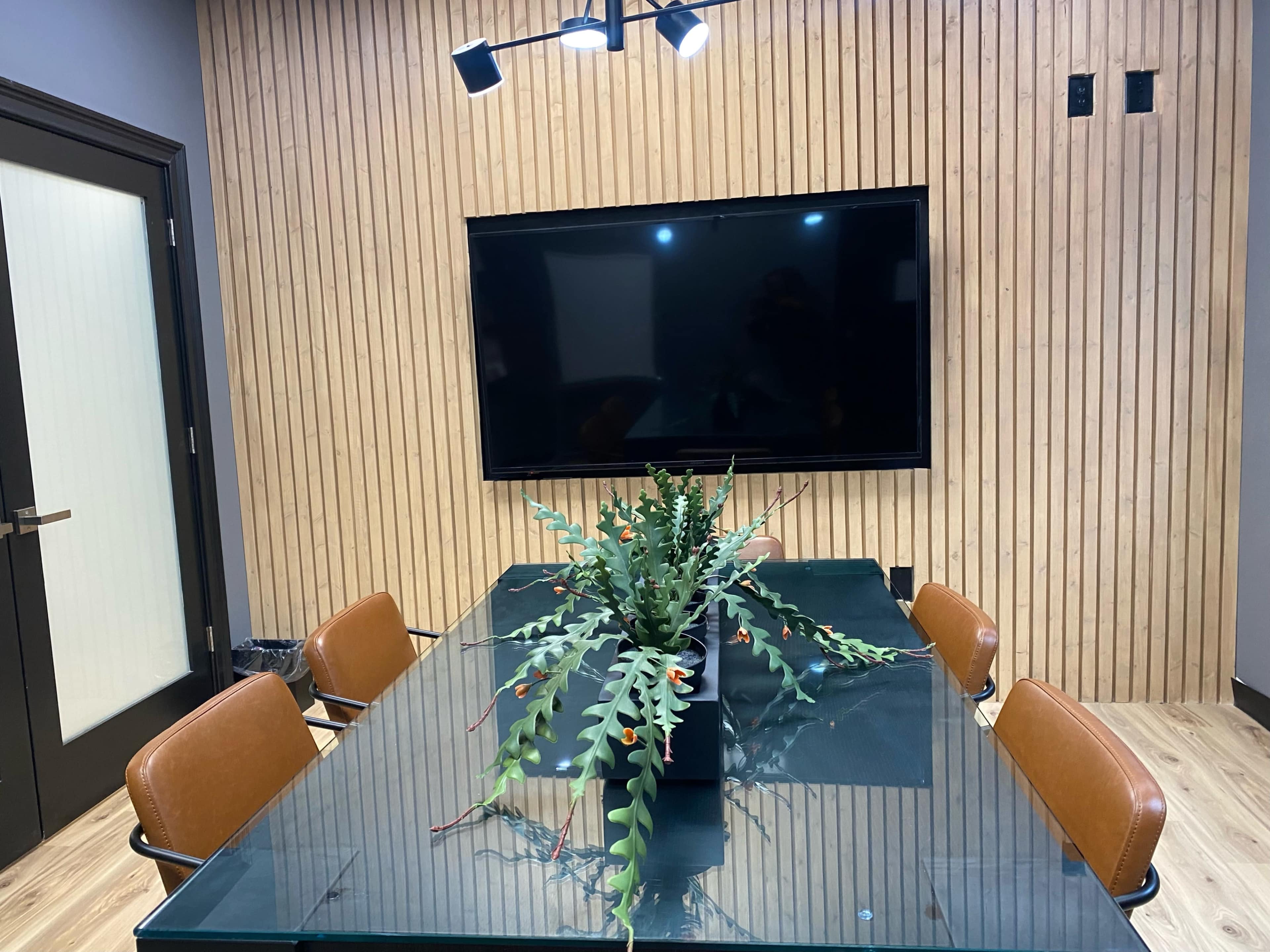 A conference room features a glass table surrounded by brown chairs, with a plant centerpiece and a large black screen mounted on a wooden wall.