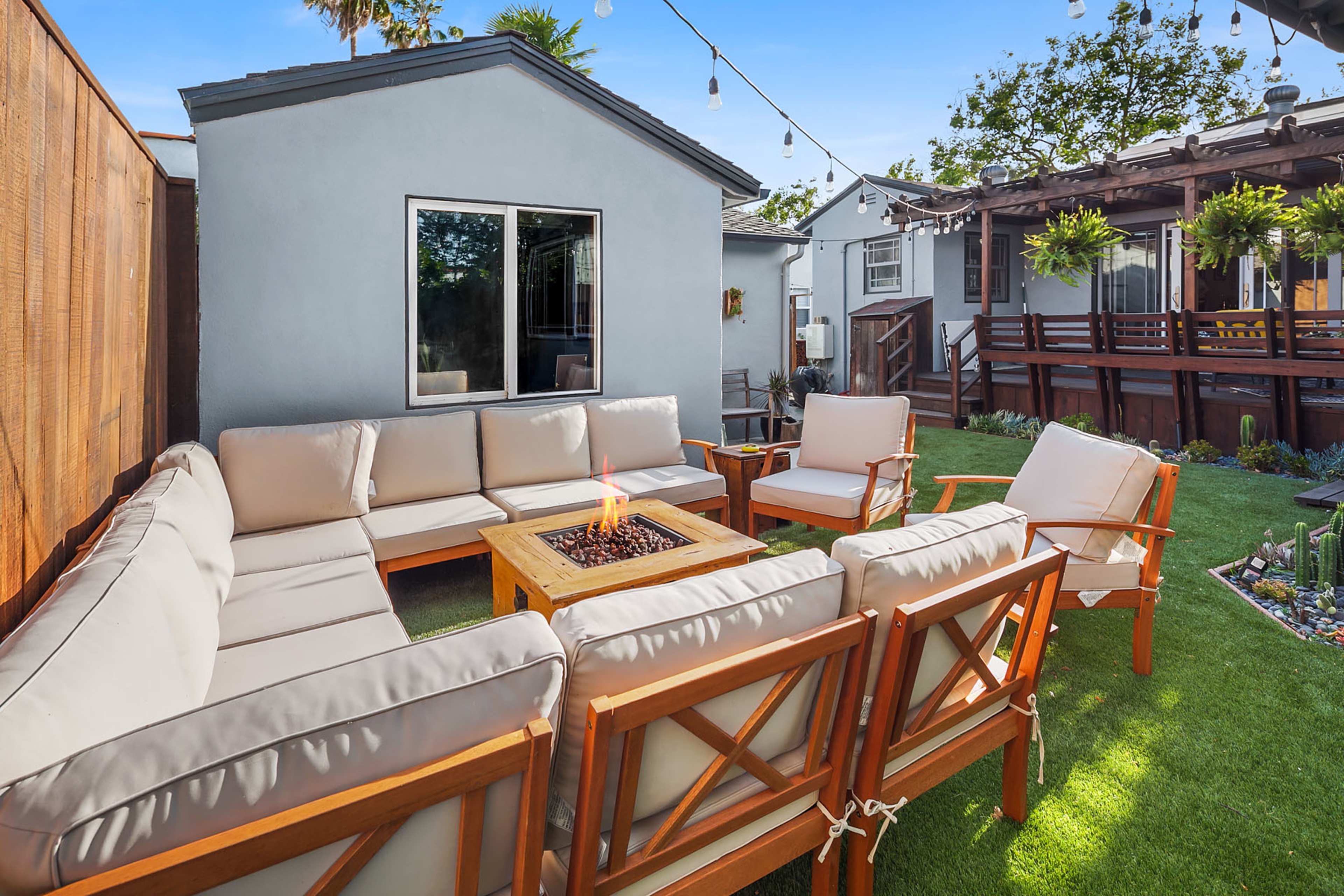 An outdoor seating area featuring a sectional sofa, a fire pit table, and several armchairs surrounded by green grass and string lights.