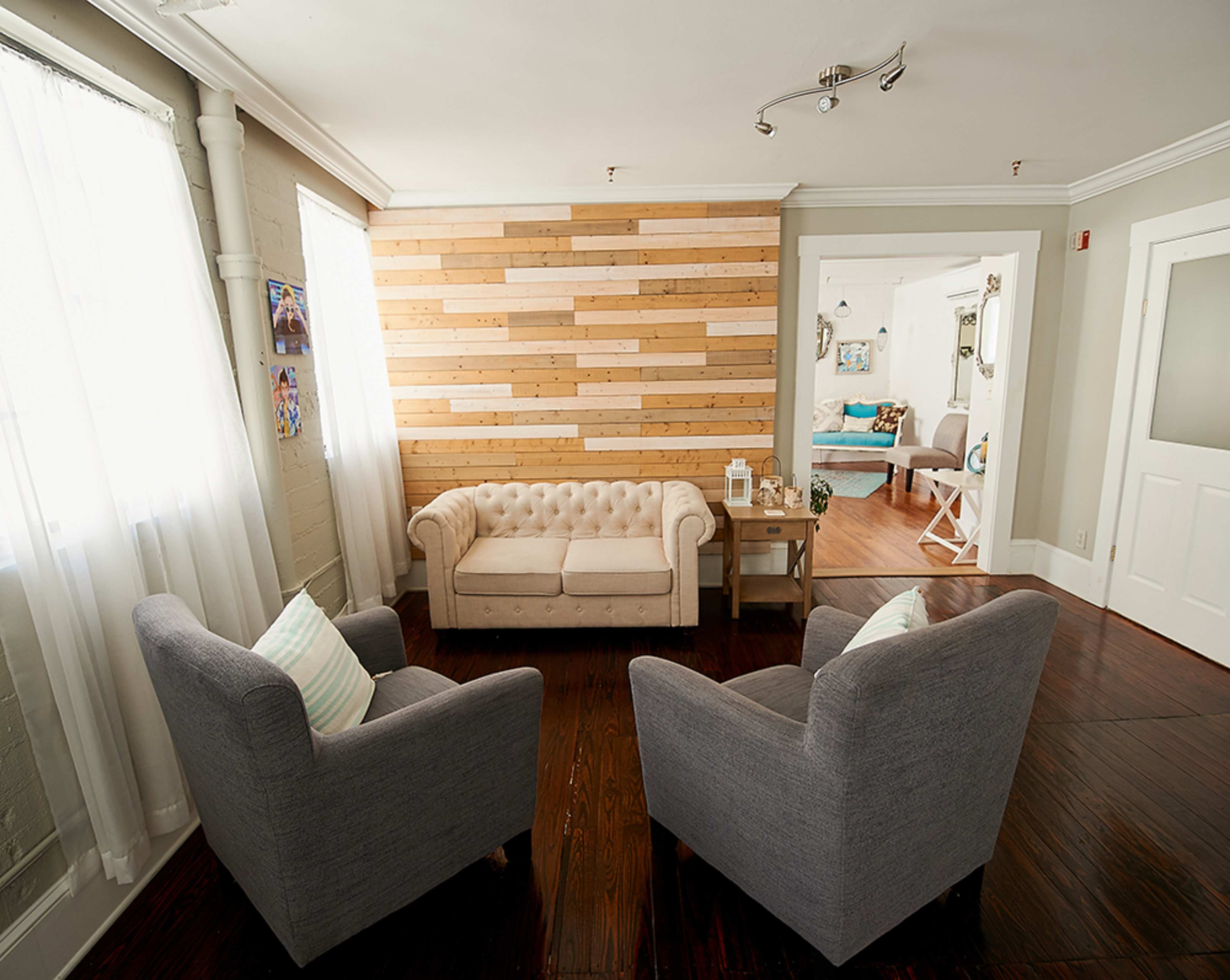 A living room features a chesterfield sofa and two gray armchairs, with a wooden accent wall and large windows letting in natural light.