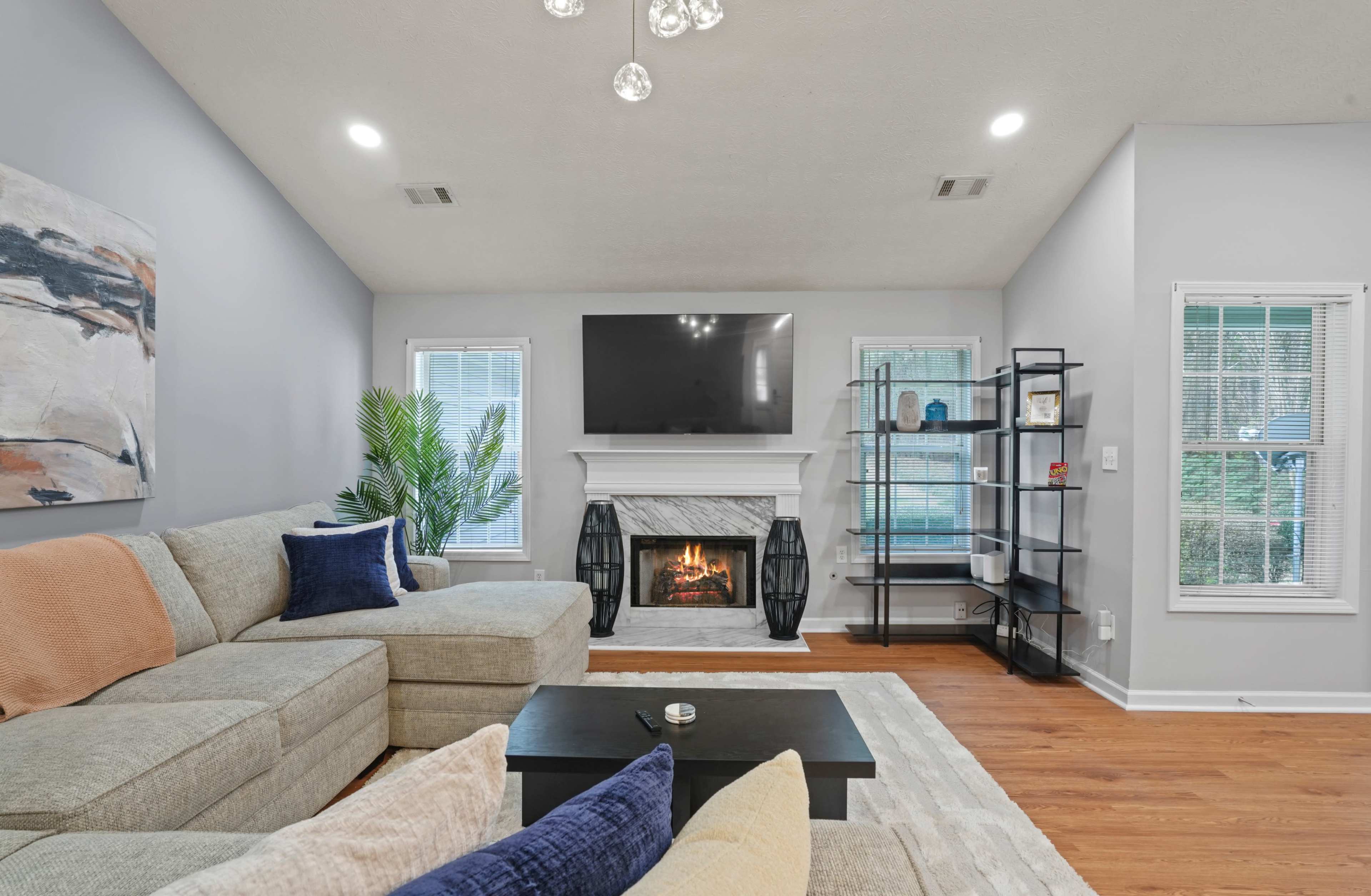 The living room features a sectional sofa, a modern fireplace with a television above it, and a corner shelf next to large windows.
