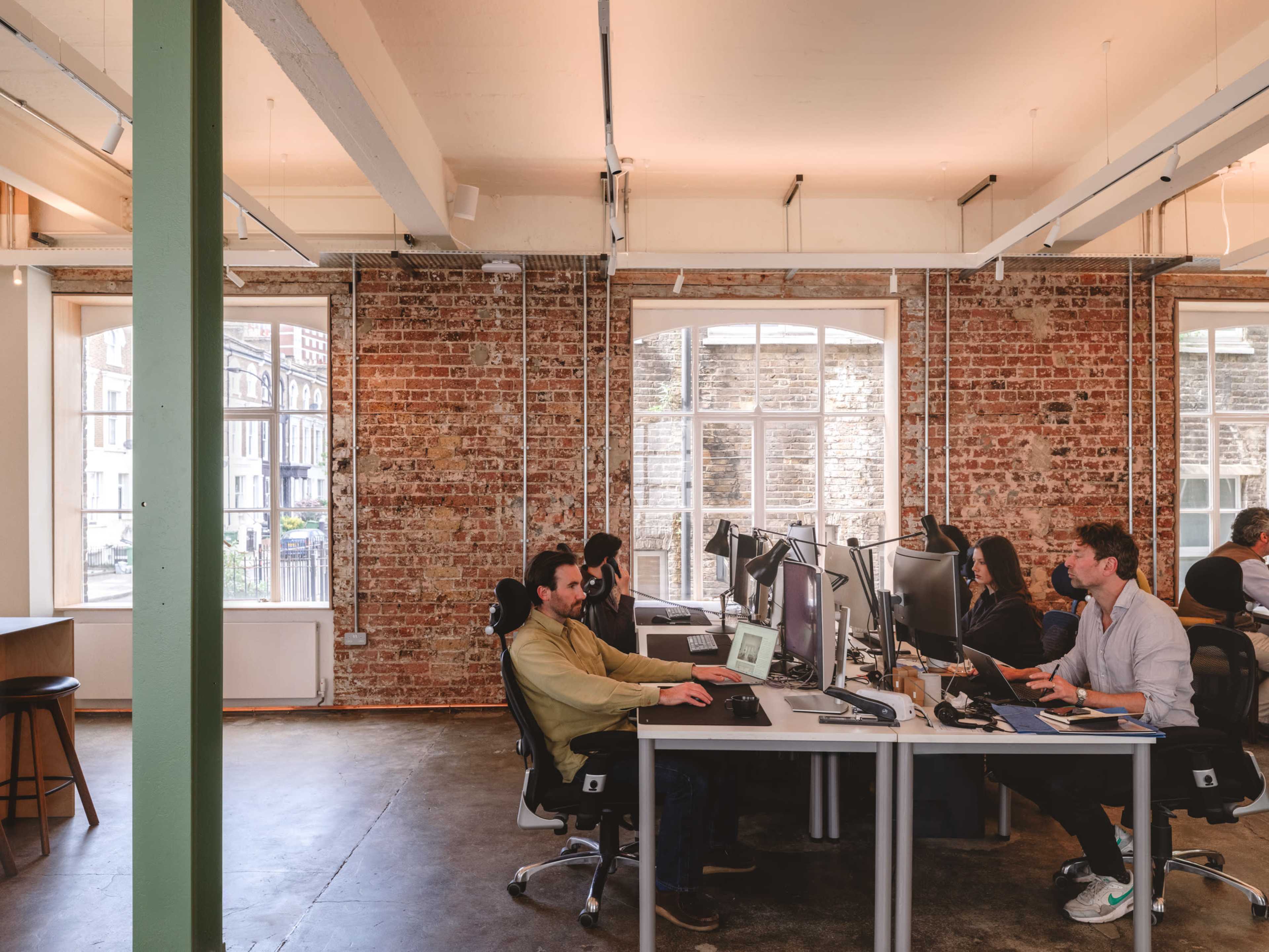 A group of people are working at desks in an office with exposed brick walls and large windows.