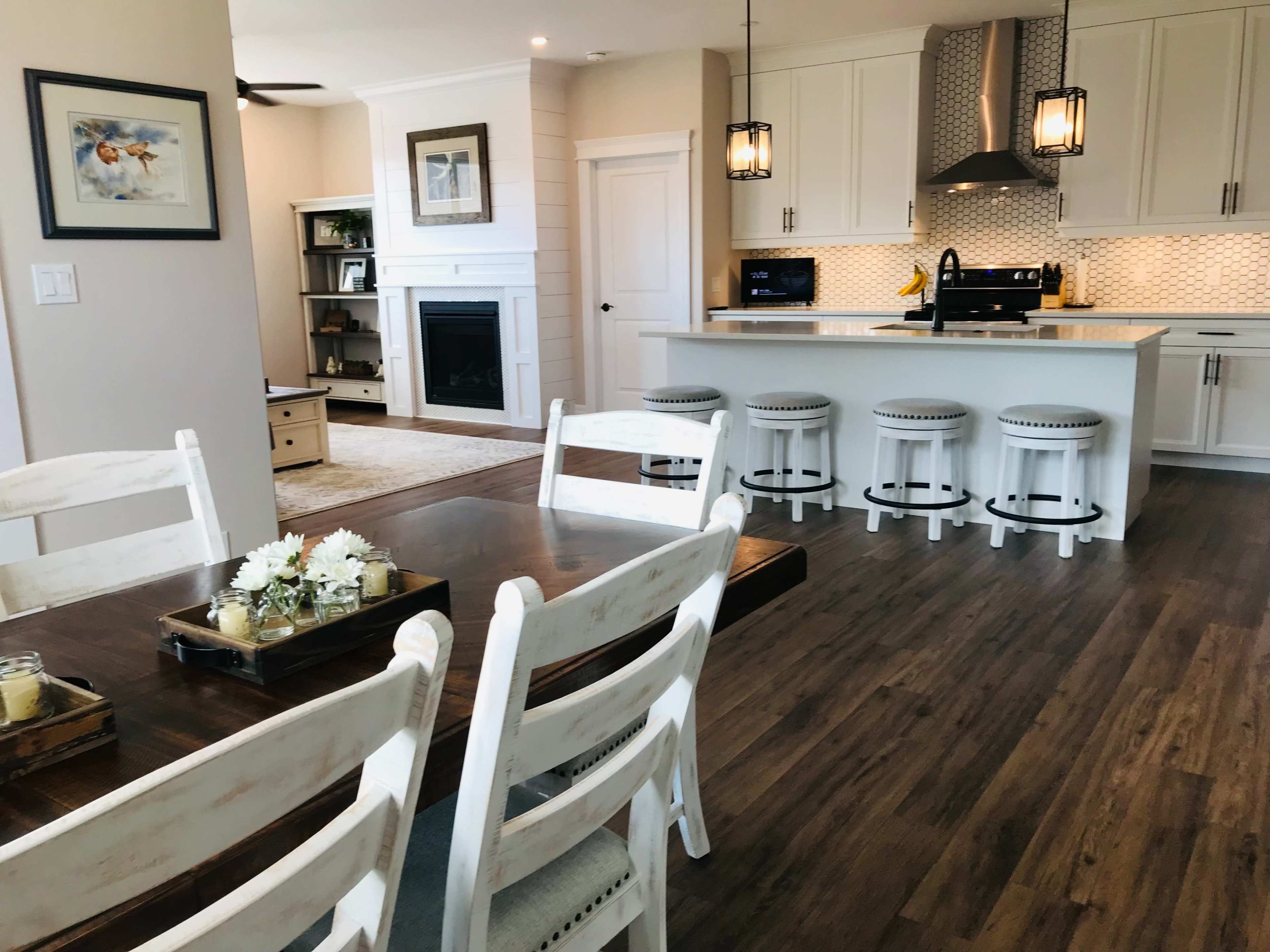 A spacious kitchen and dining area features a wooden table, white chairs, and a modern kitchen with bar stools, pendant lights, and a fireplace in the background.