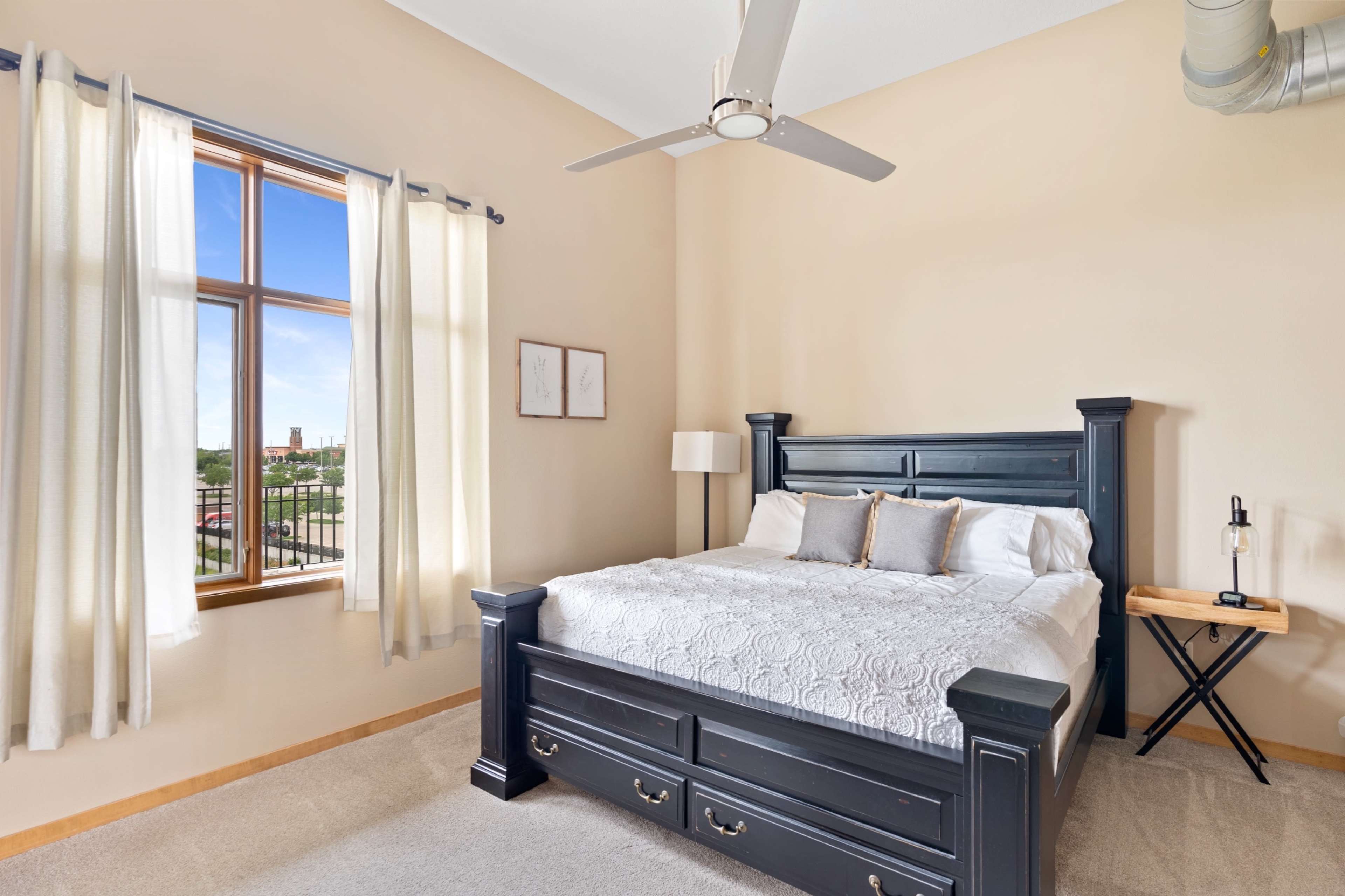 A well-lit bedroom with a dark wooden bed frame, a nightstand, and large windows allowing natural light to enter.
