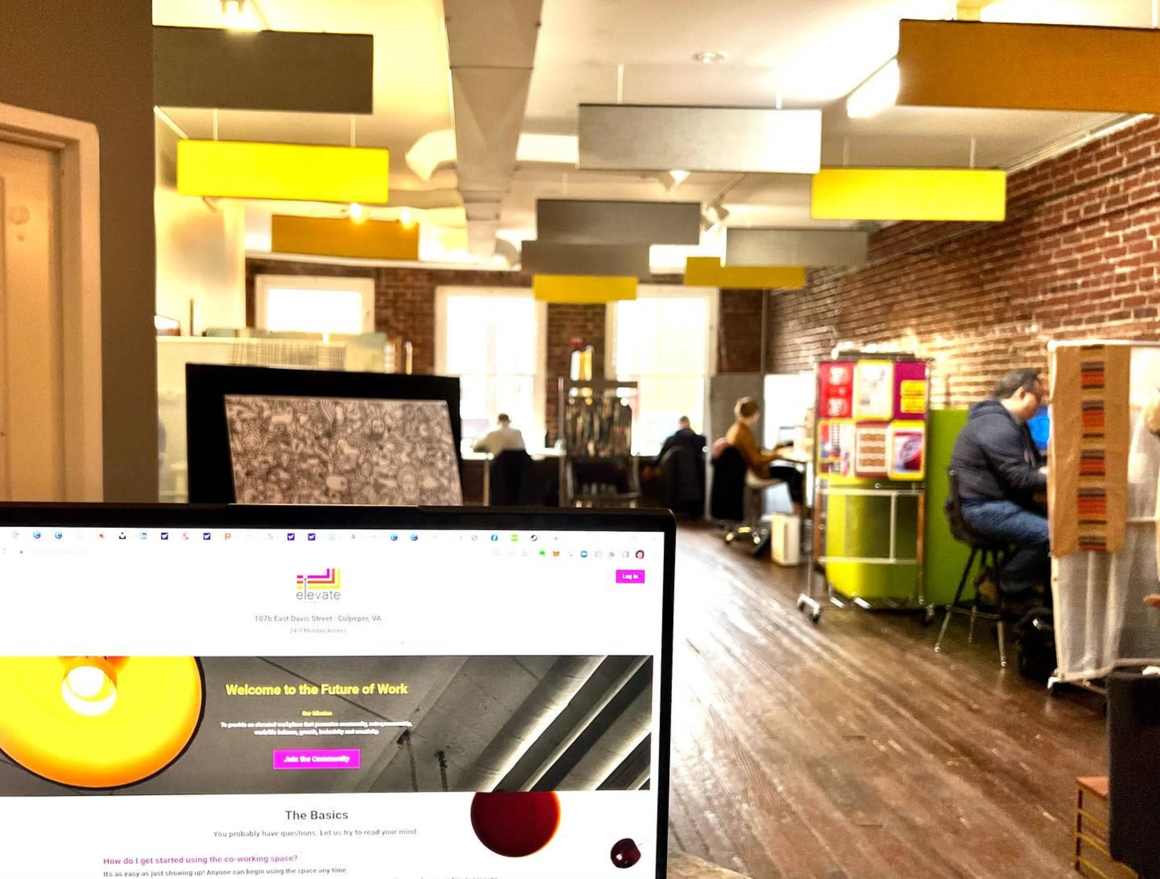 The image shows a workspace with wooden flooring and rows of desks occupied by people, with yellow light fixtures hanging from the ceiling.