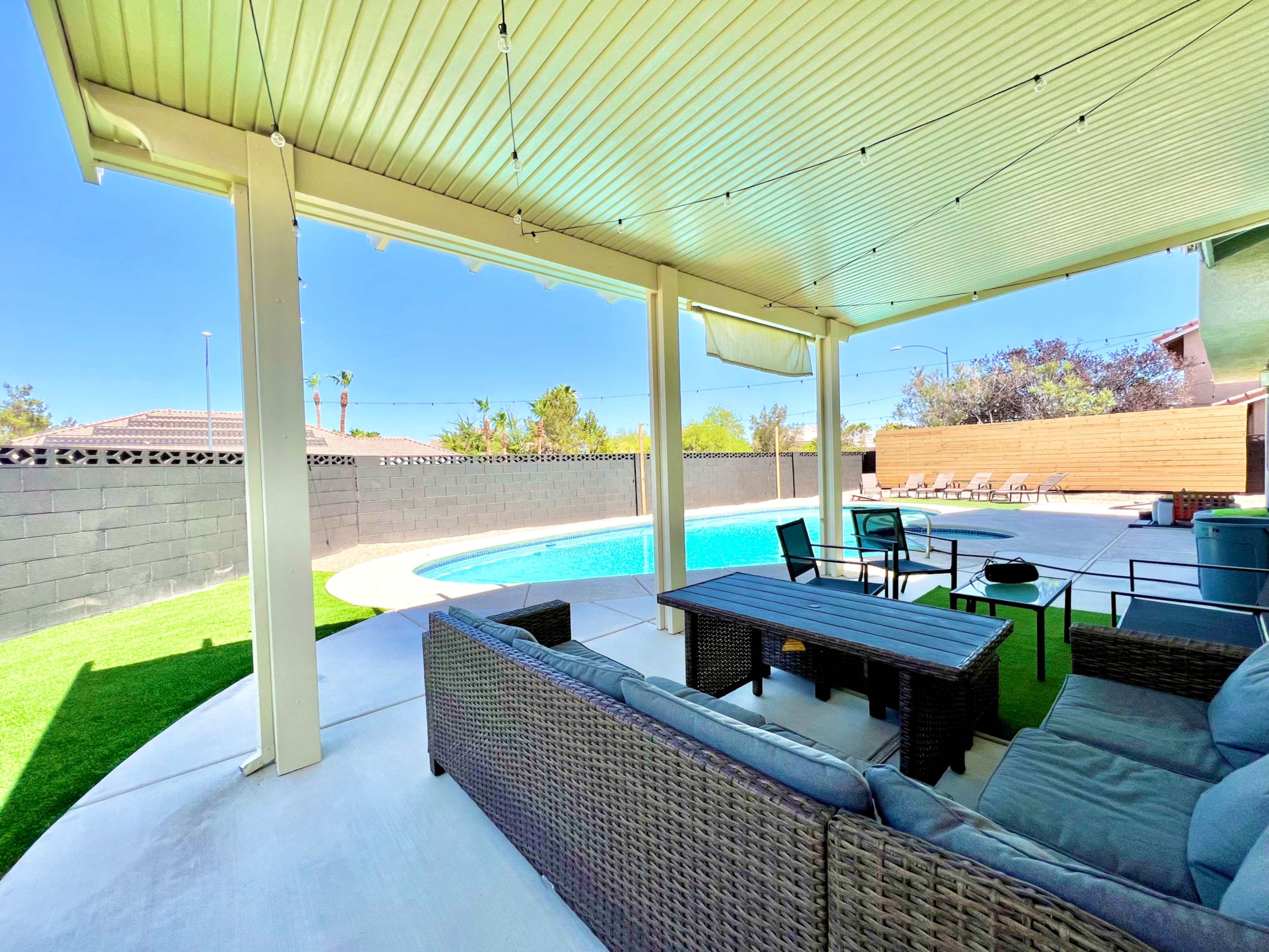 A covered patio area with seating and a table, overlooking a swimming pool surrounded by a landscaped yard.