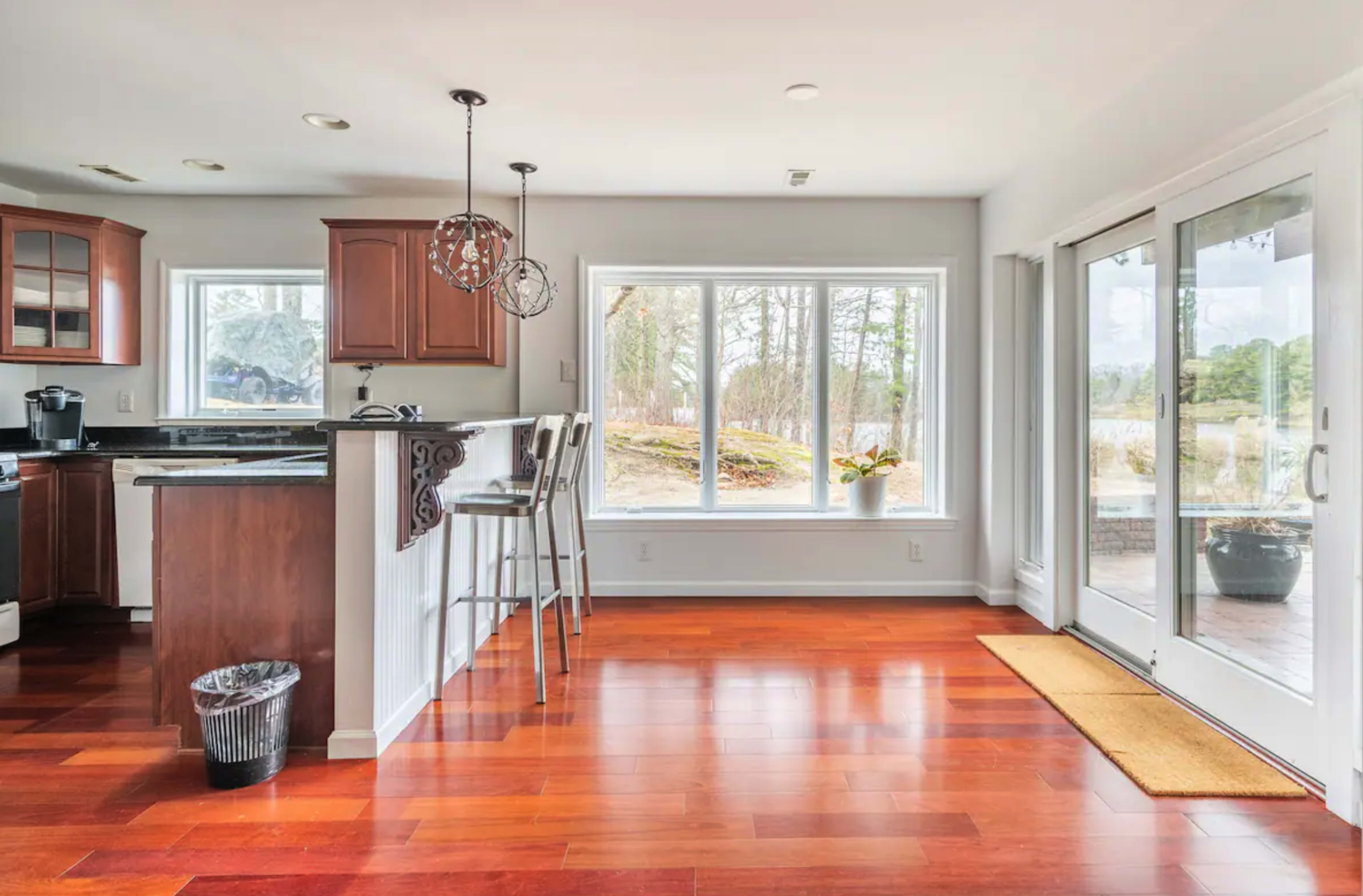 A modern kitchen features wooden cabinets, a bar with stools, and large windows overlooking a wooded area.