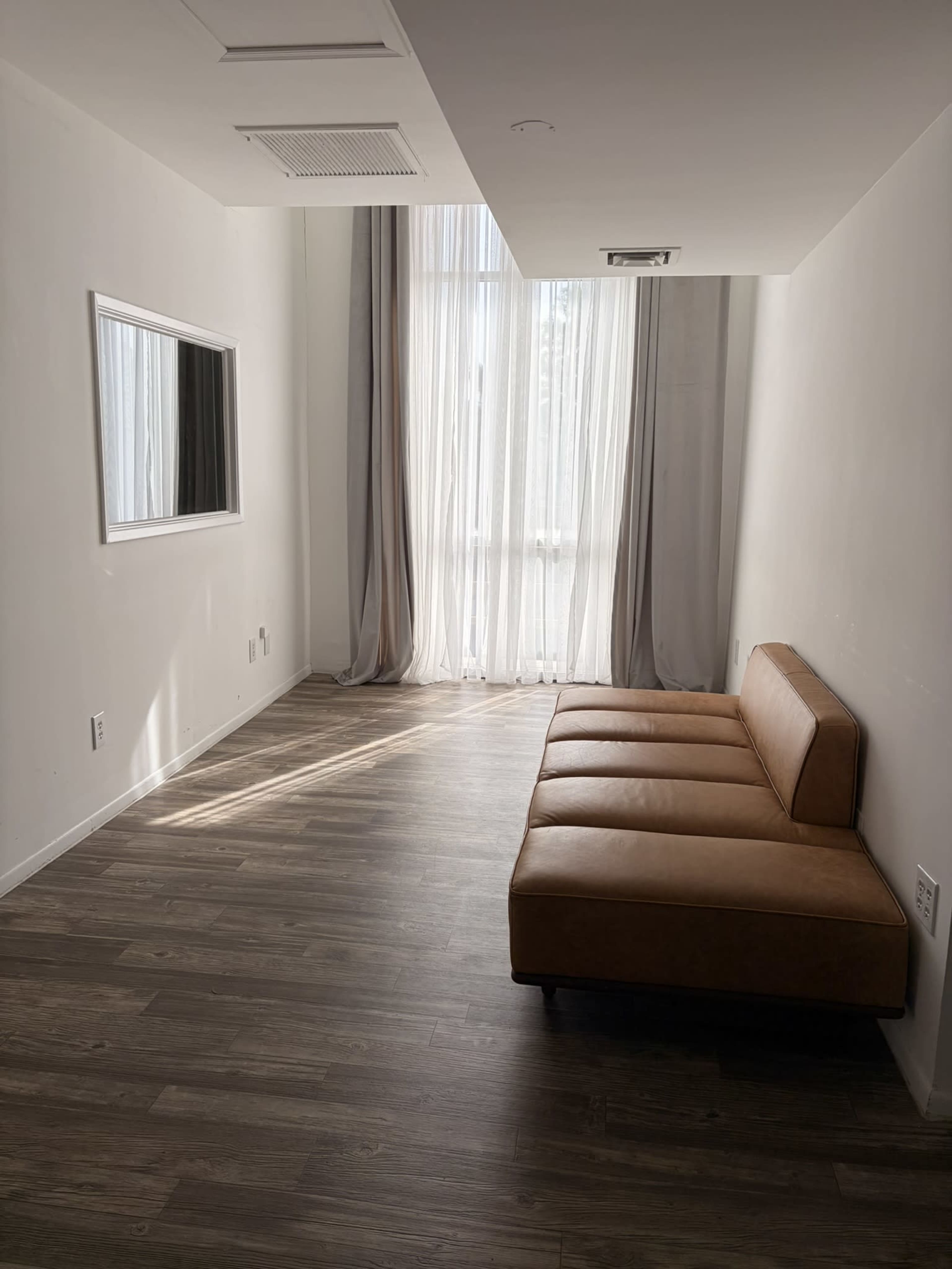 The image shows a minimalist room featuring a tan, upholstered sofa, a large window with sheer curtains, and a wall-mounted mirror.