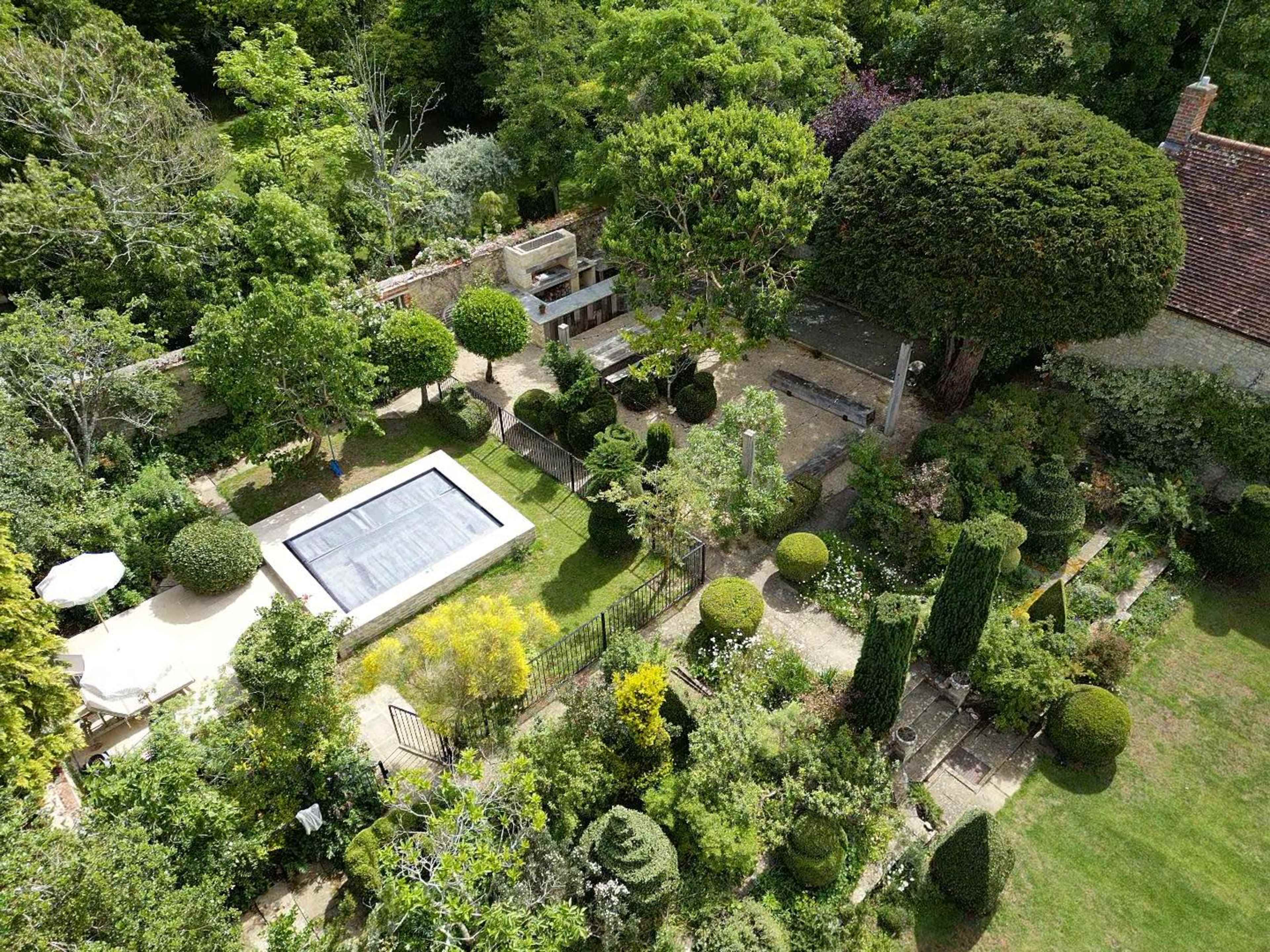 The image shows a landscaped garden with a swimming pool, various plants, and neatly trimmed hedges surrounded by trees.