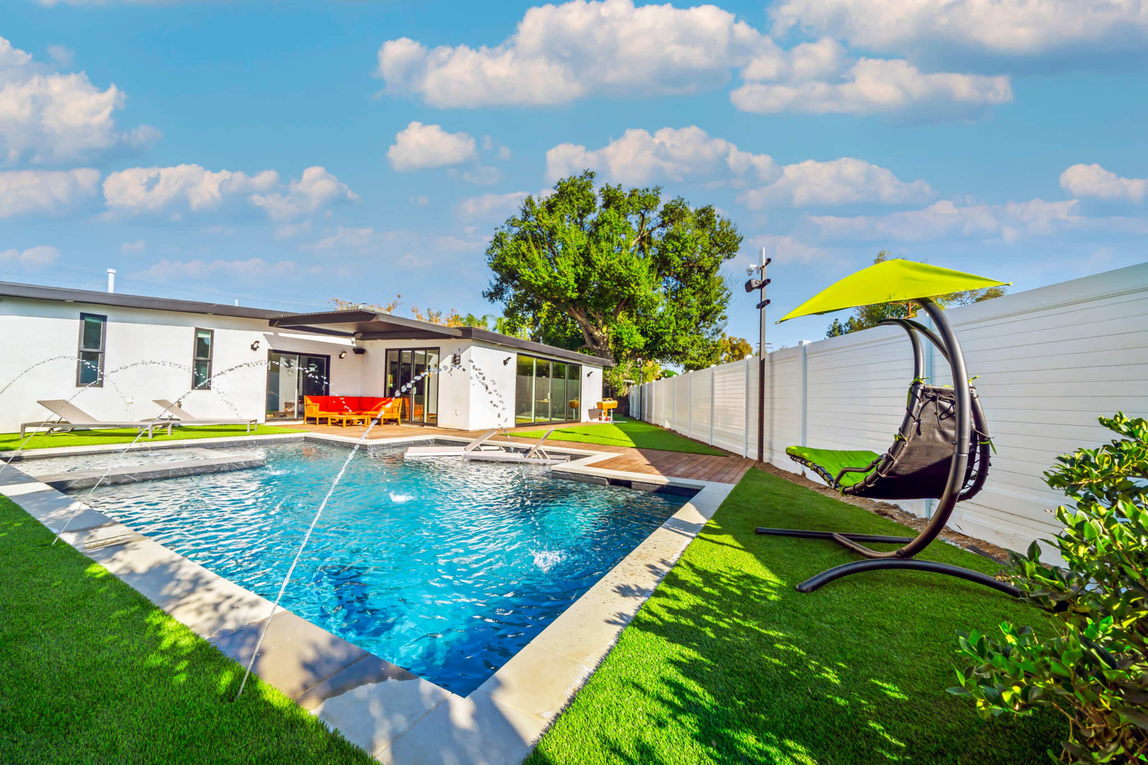 A modern backyard featuring a swimming pool with water fountains, a hanging chair, and a house in the background, surrounded by green grass and white fencing.