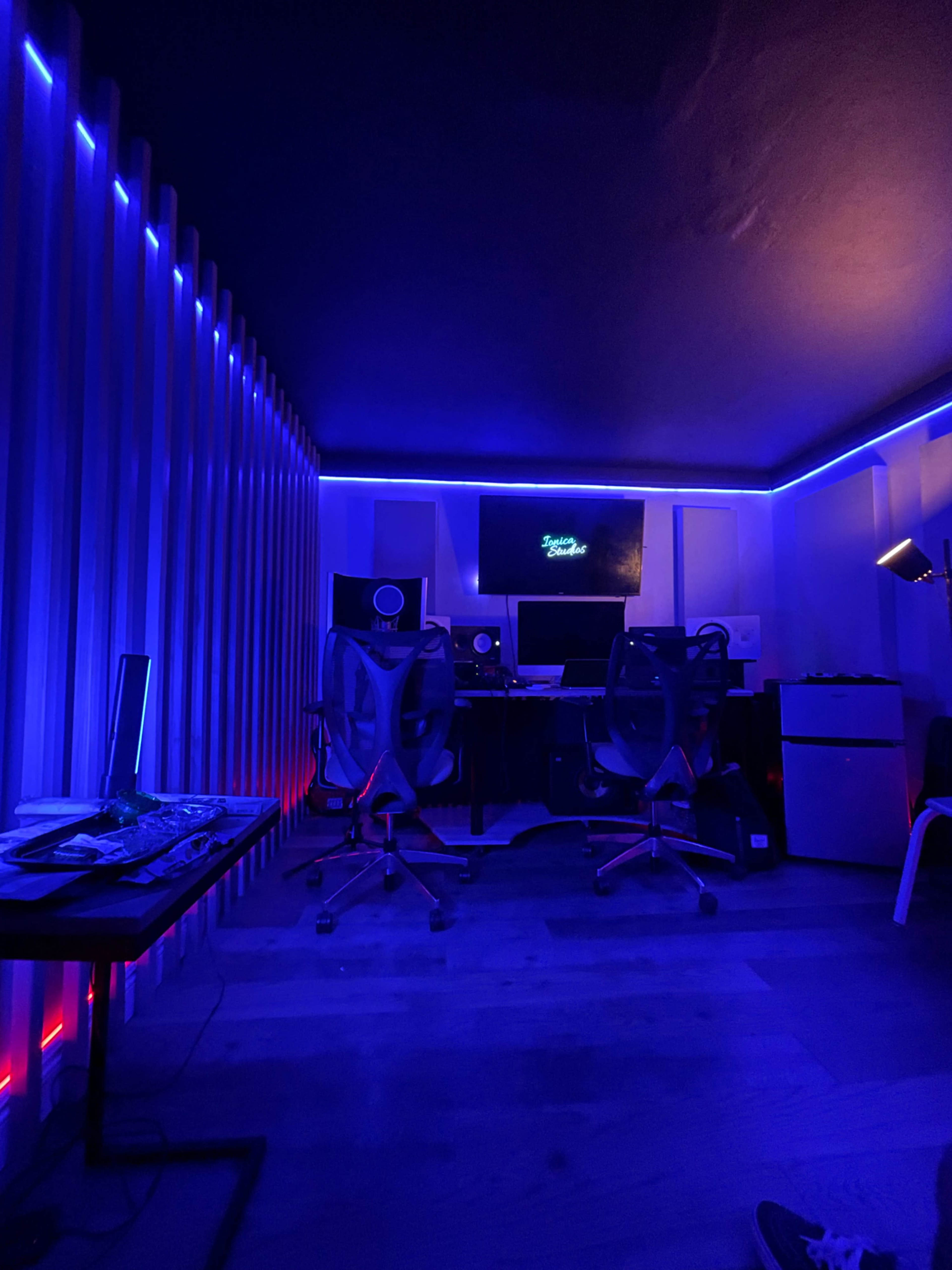 The image displays a music studio with blue LED lighting, featuring two office chairs and a monitor in front of a sound setup against illuminated walls.